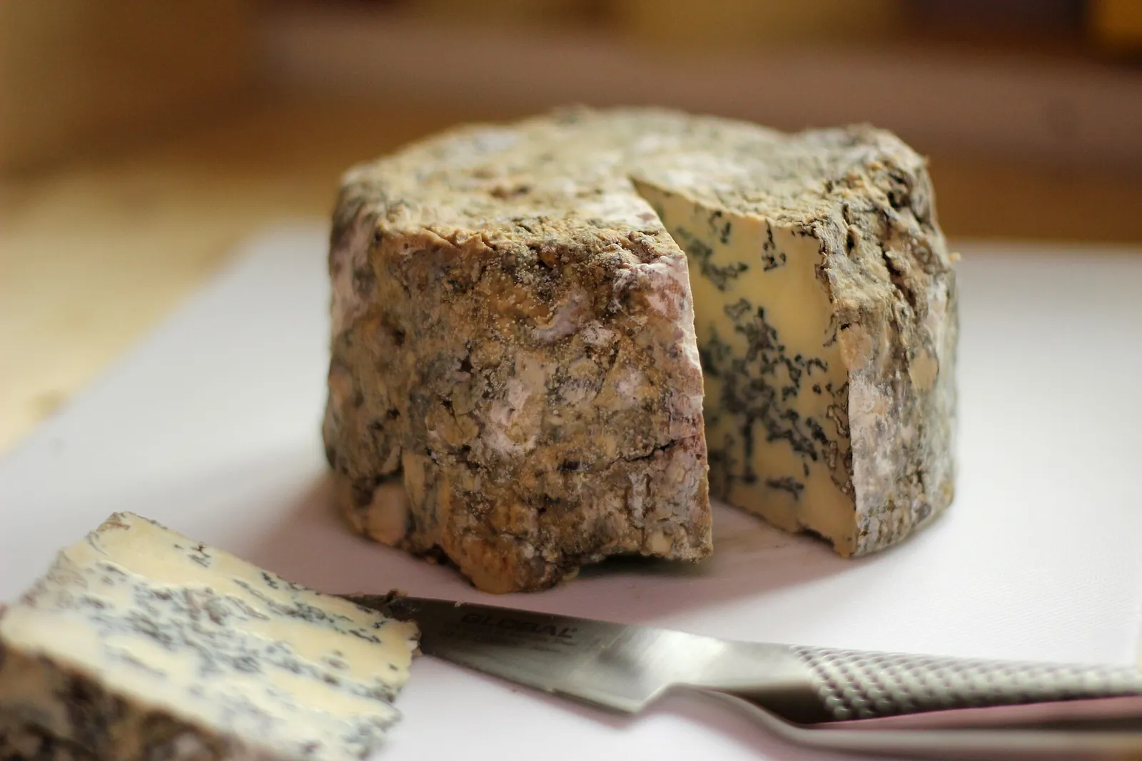A rustic blue cheese with a gray-brown natural rind and a wedge cut away showing blue-green veins, with a cheese knife in front