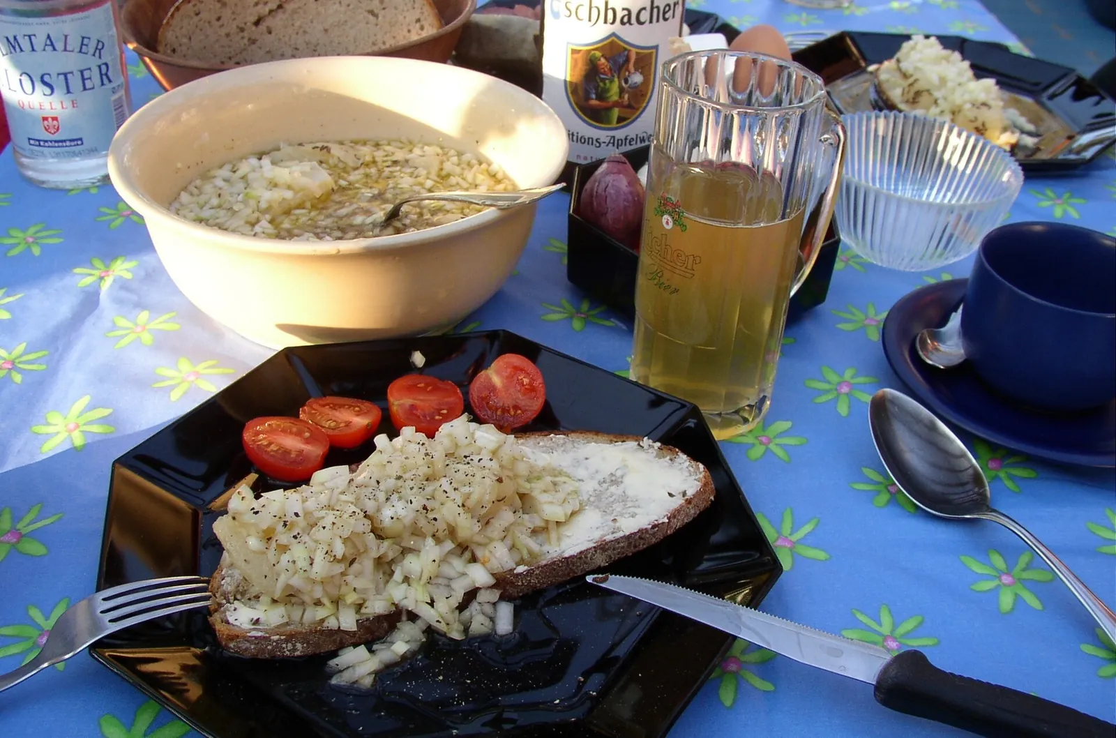 Handkäse cheese with chopped onions on dark bread beside a pitcher of golden apple wine, cherry tomatoes and a bowl of marinated cheese.