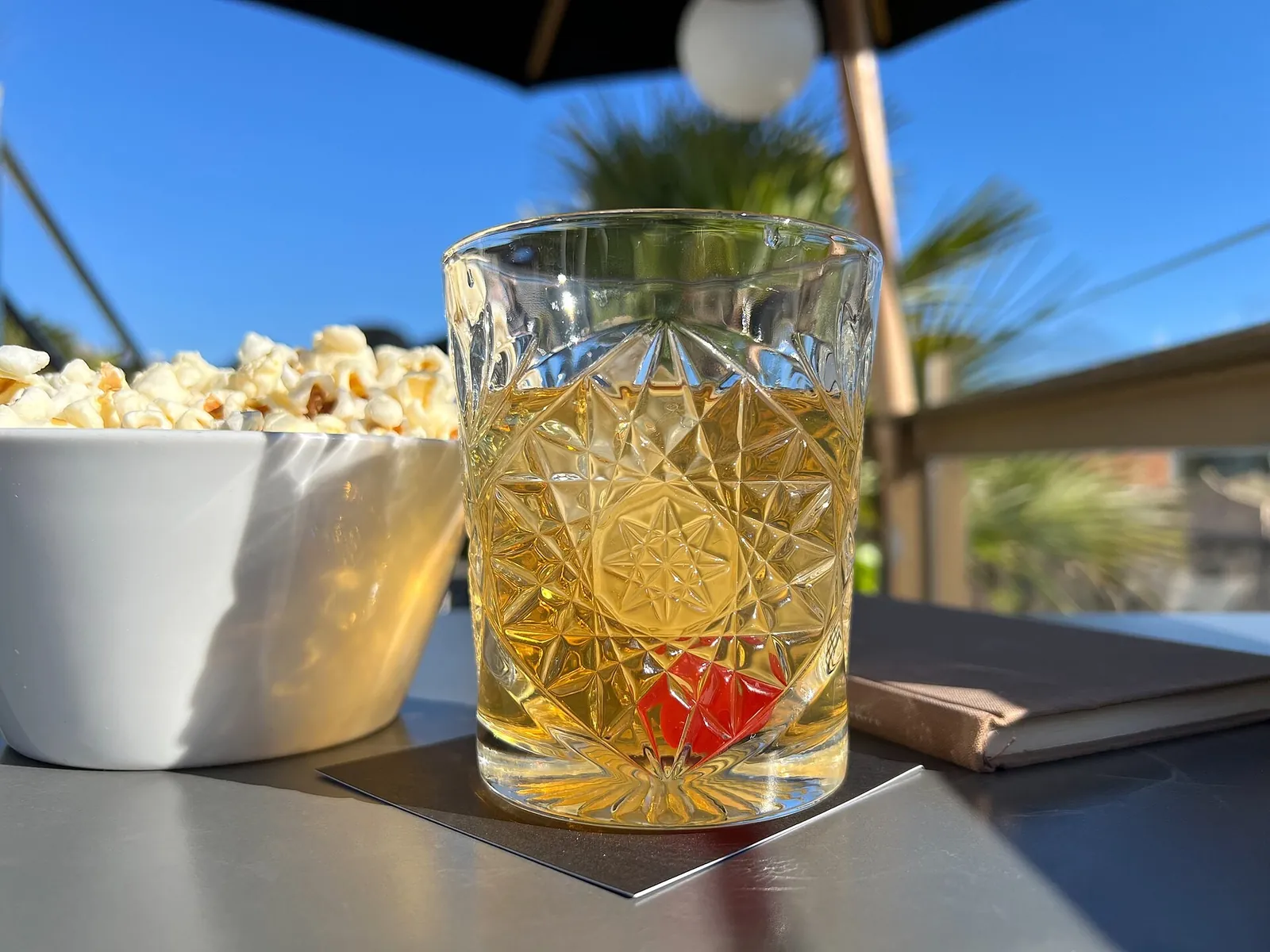 A cut-glass tumbler of golden Old Fashioned with a red cherry, beside a white bowl of popcorn on a sunny terrace table.