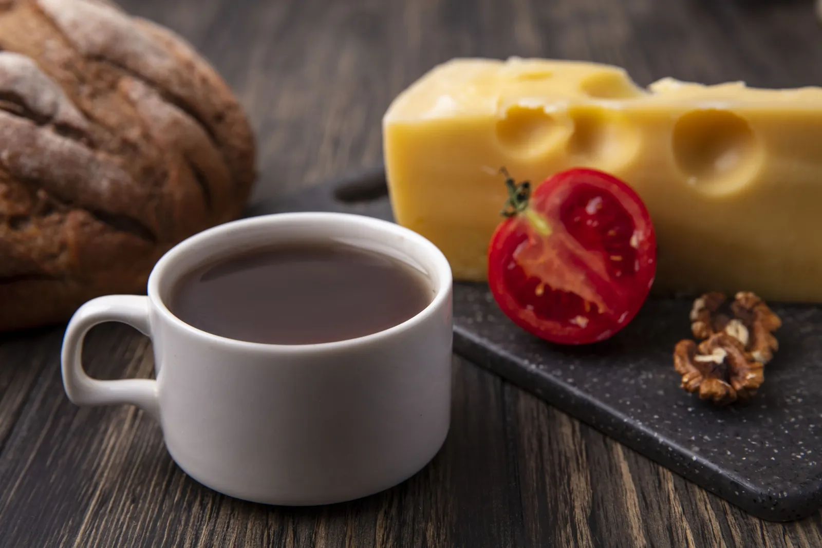 A white cup of coffee in front of a block of holey yellow cheese, half a tomato and walnuts on a board, with a dark loaf behind.