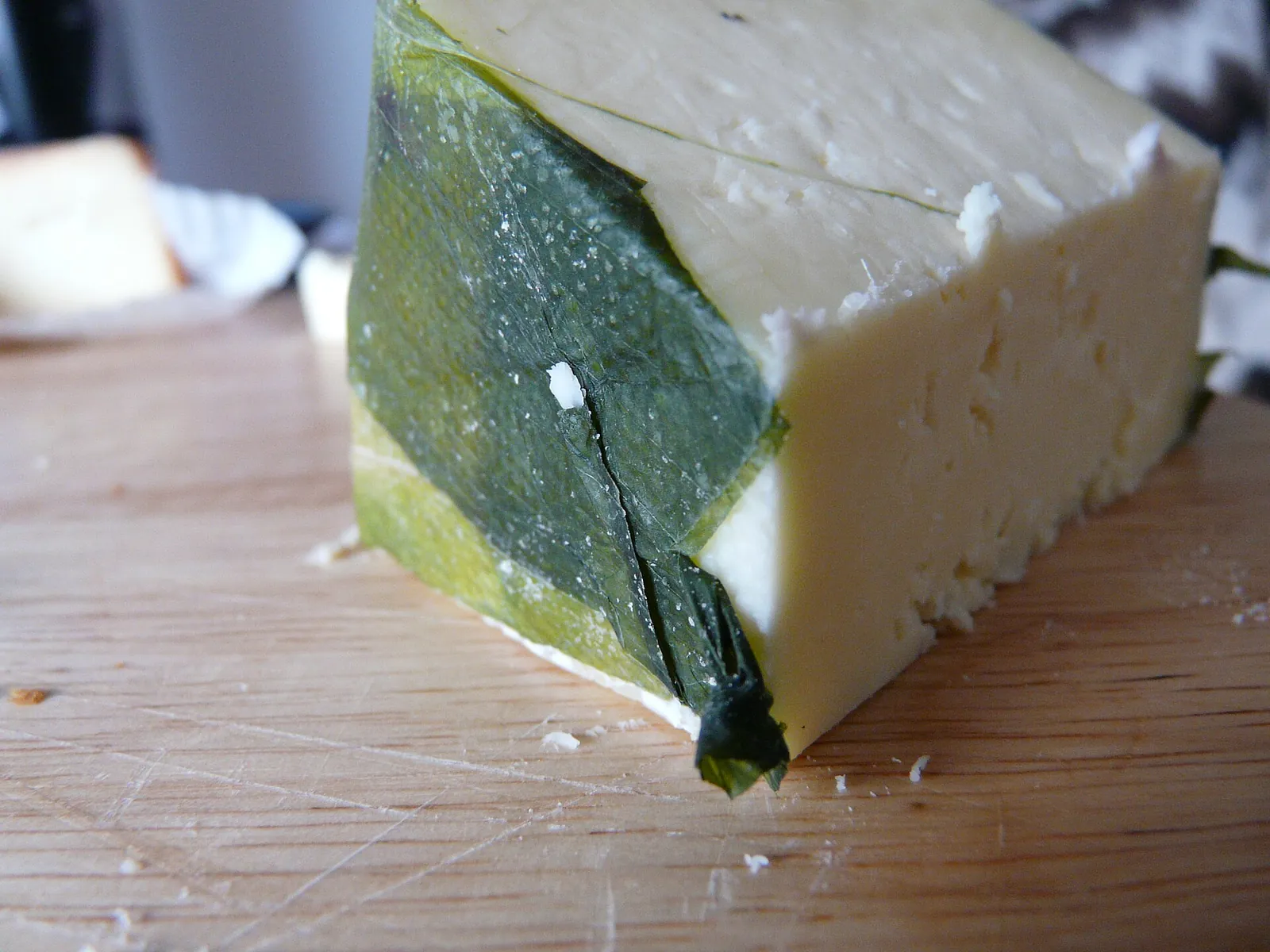 A wedge of Yarg wrapped in dark green leaves, with pale crumbly cheese showing, on a wooden board