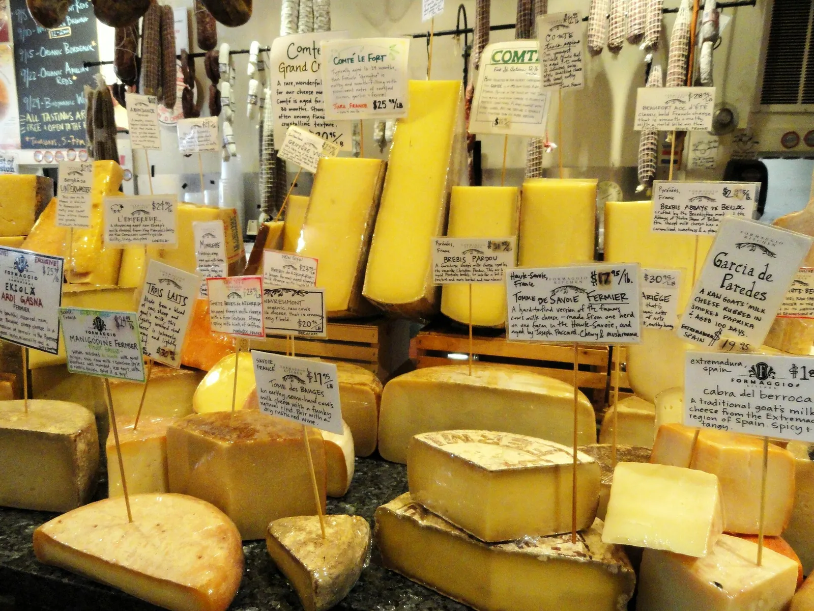 A cheese counter with wedges of Comté, Gouda and other cheeses under handwritten label stakes