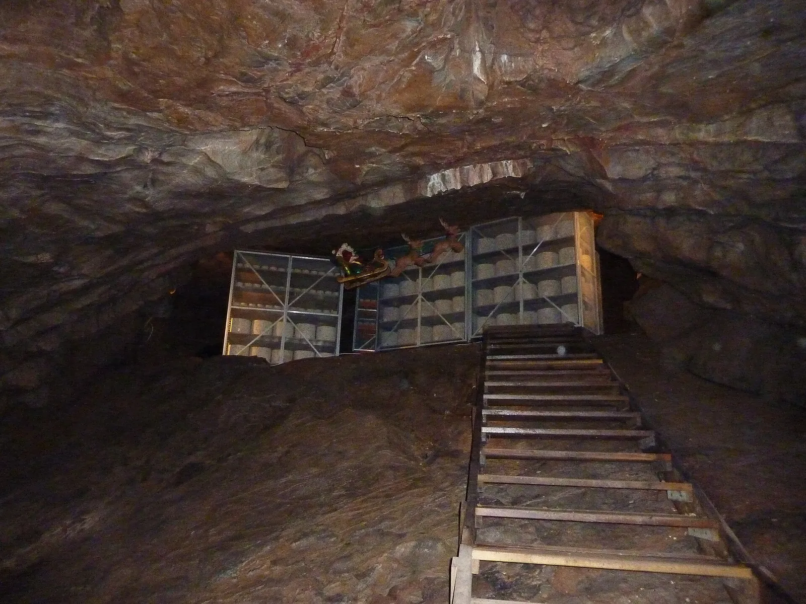 A dark rock cave at Cheddar Gorge in Somerset, England, with wooden racks of maturing cheese beyond a short flight of steps.