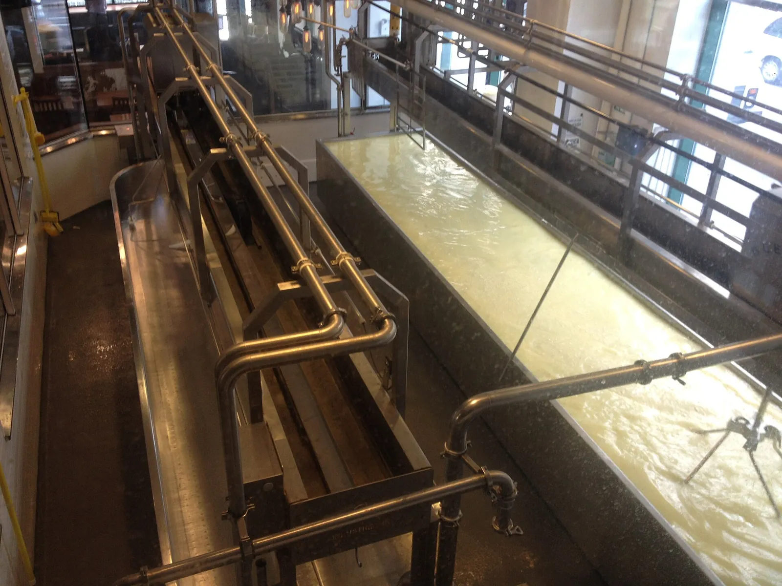A long stainless steel vat of fresh cheese curds and whey, with pipes and railings above, at a cheesemaker in New York City.