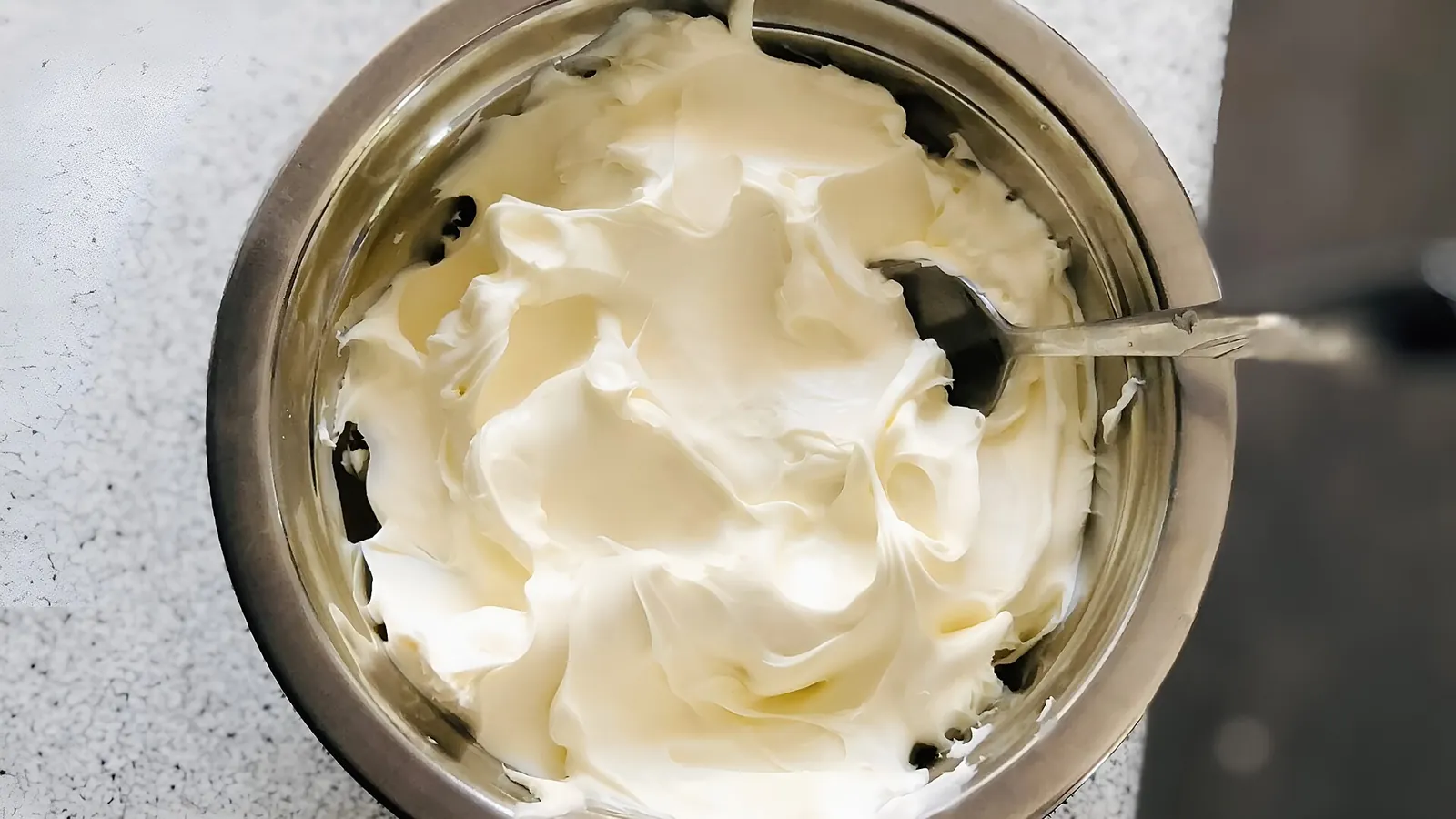 A steel bowl of thick, pale homemade mascarpone with a spoon resting in it on a stone counter