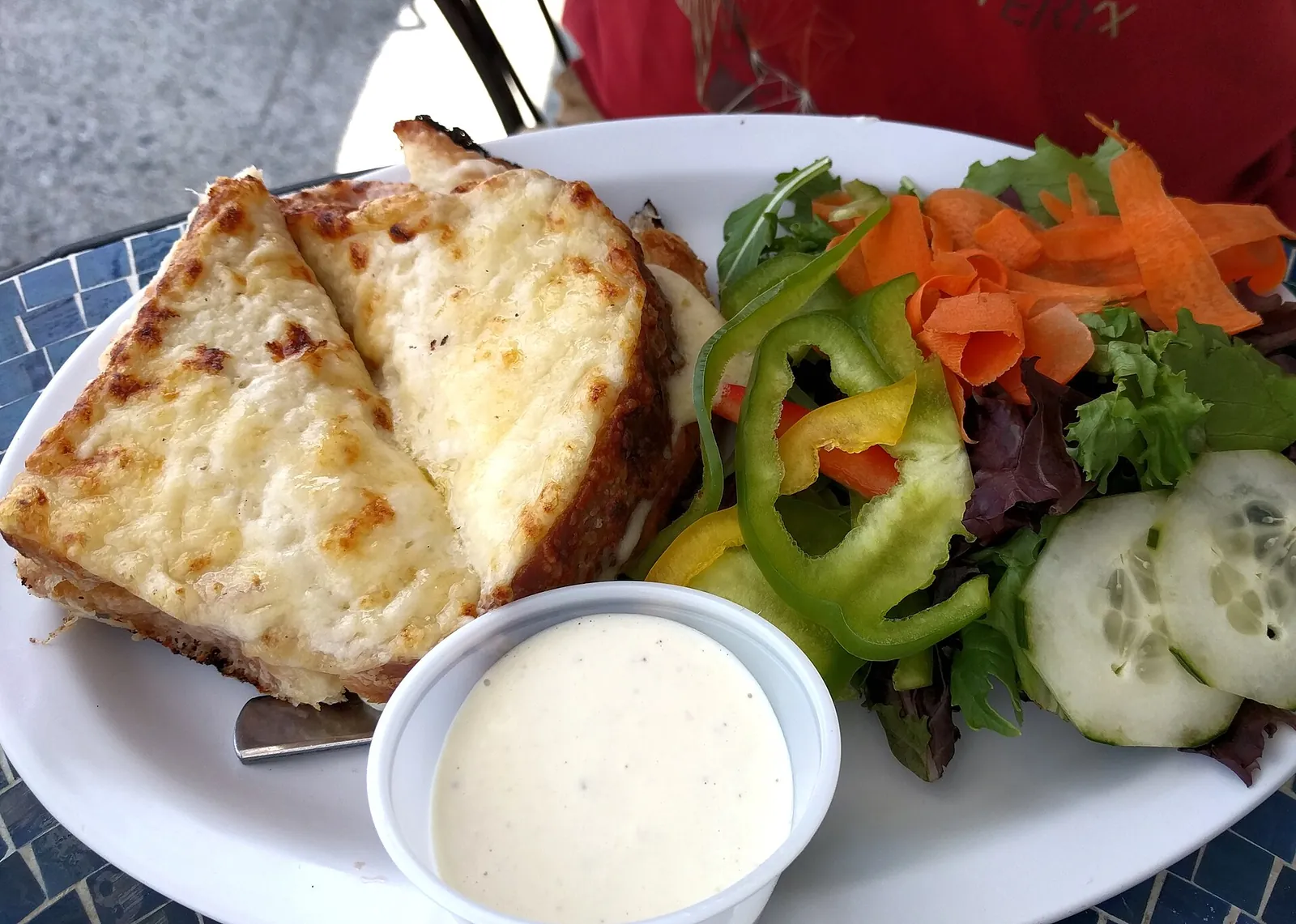 A croque monsieur with a browned, bubbling cheese top, served on a plate with a green salad and a small dish of white sauce.
