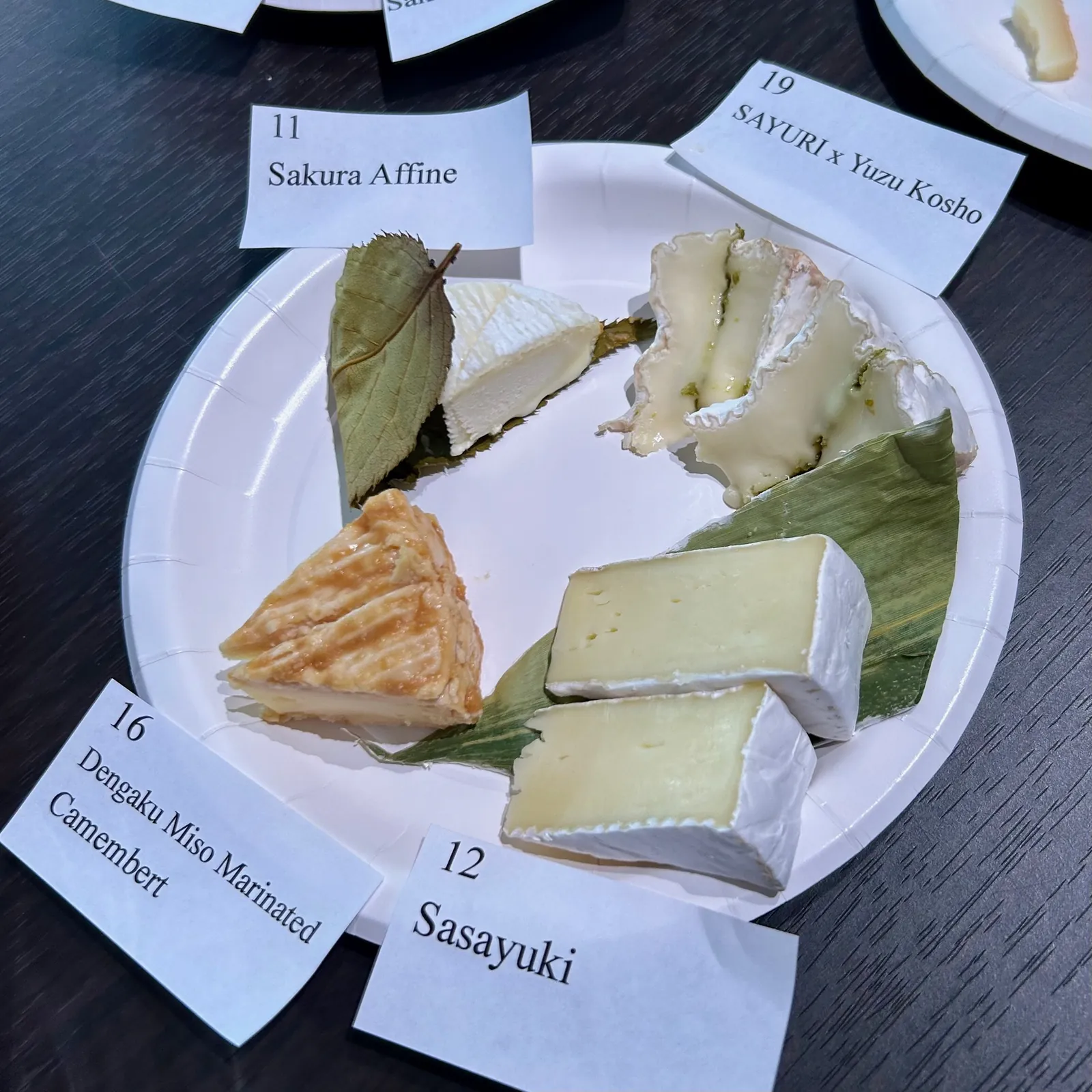 A paper plate of Japanese cheeses at a tasting, with soft white wedges, one on a leaf, and numbered paper labels around the edge.