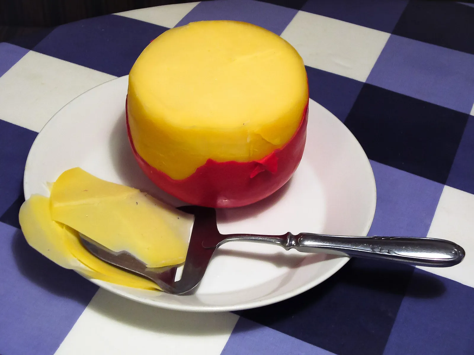 A small Edam in red wax with its yellow face cut, beside slices and a cheese slicer on a plate