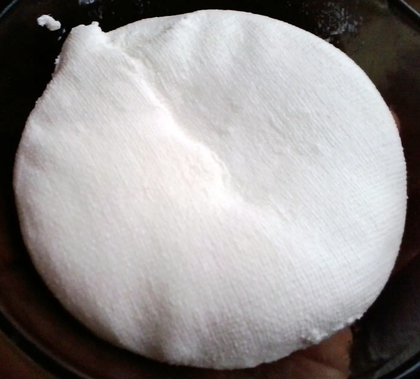 A round of white farmer cheese wrapped in cheesecloth, the weave of the cloth pressed into its surface, on a dark pan.