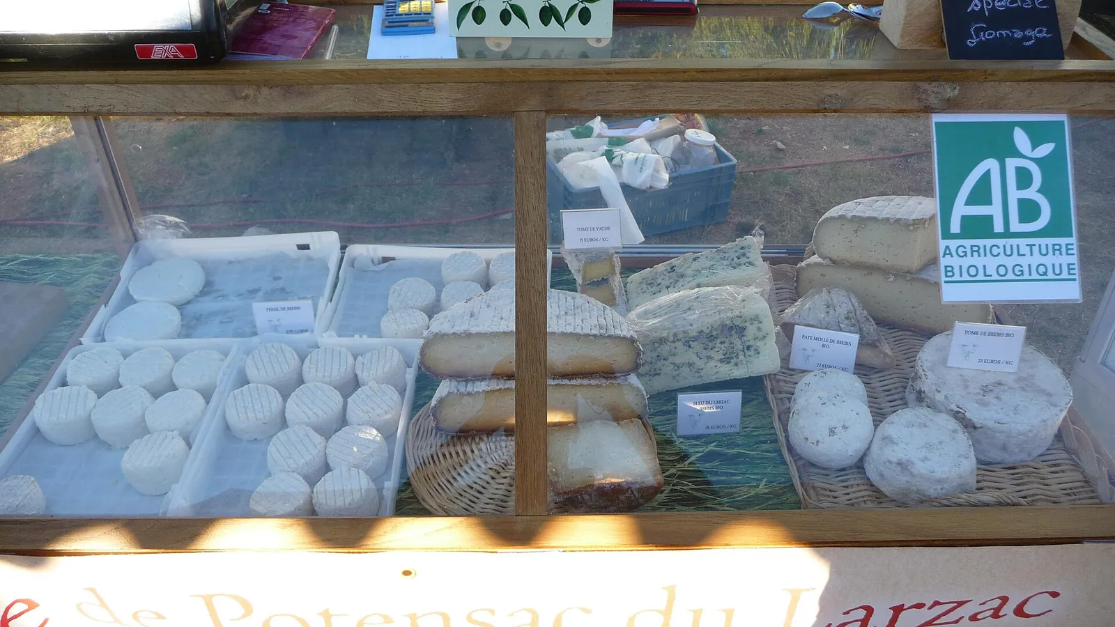 A farm shop display case of small white fresh cheese rounds and wedges of farm cheese, with a green organic farming label on the glass