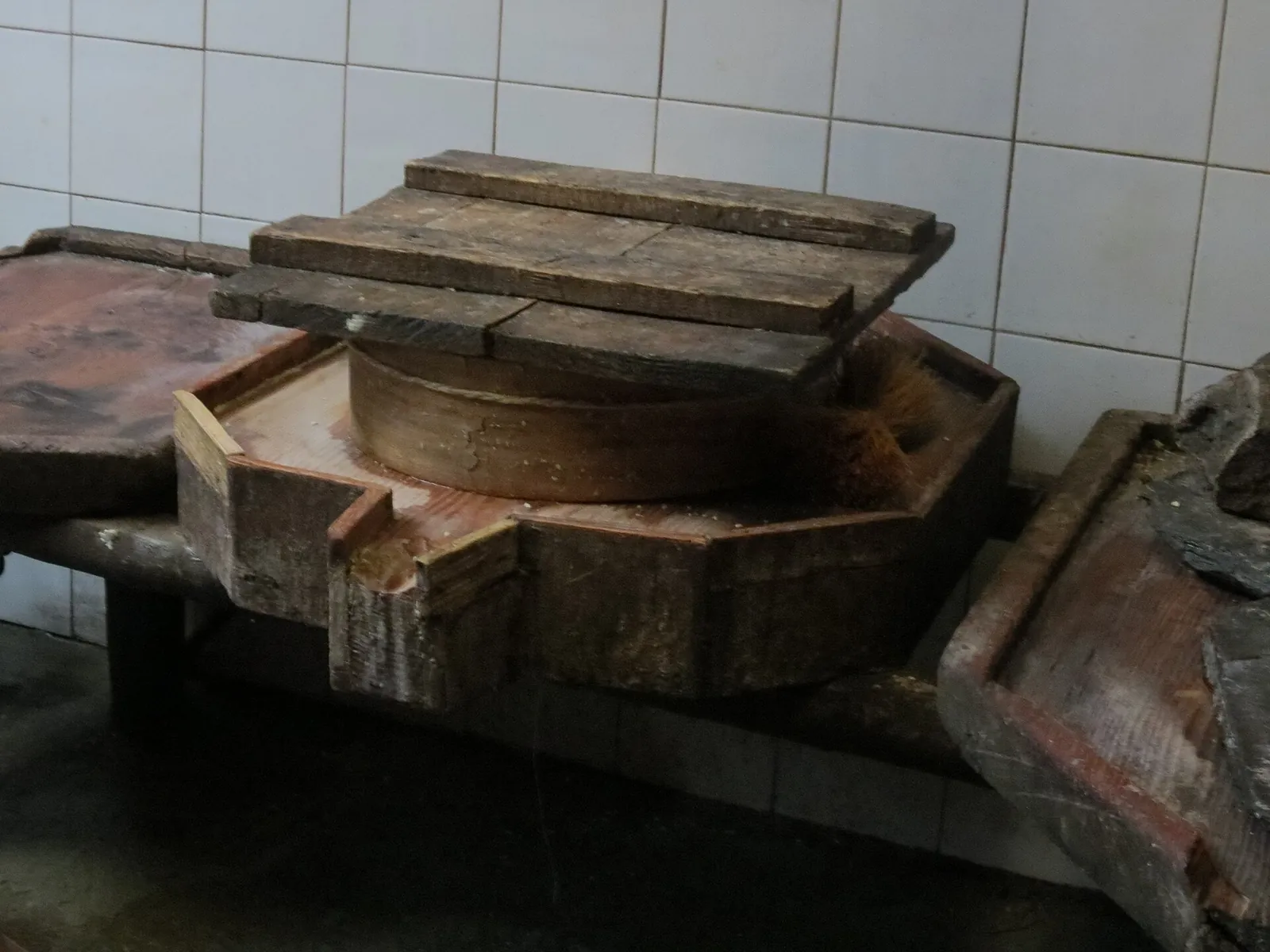 A wheel of cheese in a round wooden mold under heavy wooden boards on an old pressing table beside a tiled wall.