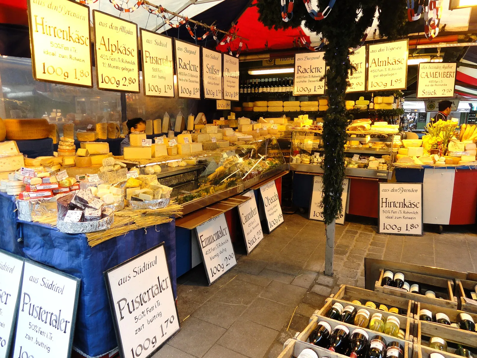 A cheese stall at the Viktualienmarkt in Munich, with wheels and wedges heaped on counters under hand-lettered signs for Alpkäse, Hirtenkäse and Raclette.
