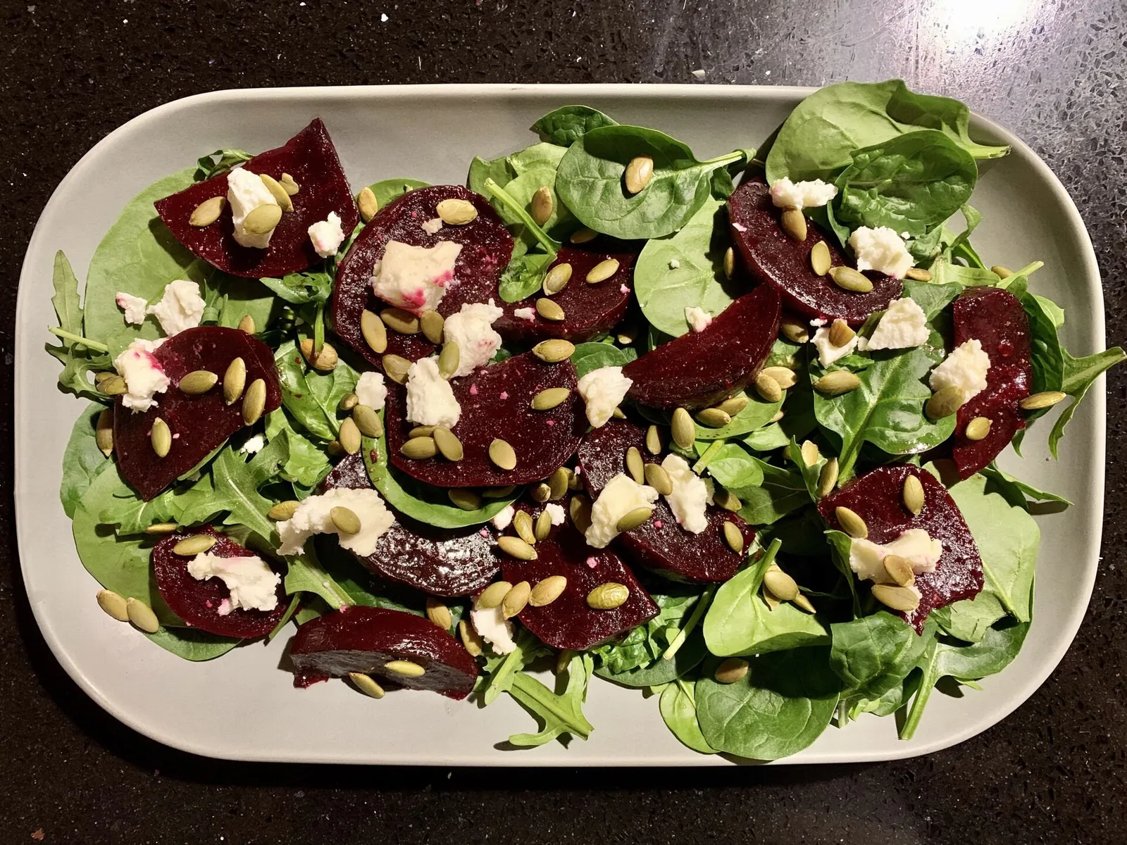 A rectangular plate of sliced roasted beets and baby spinach with crumbled goat cheese and green pumpkin seeds.