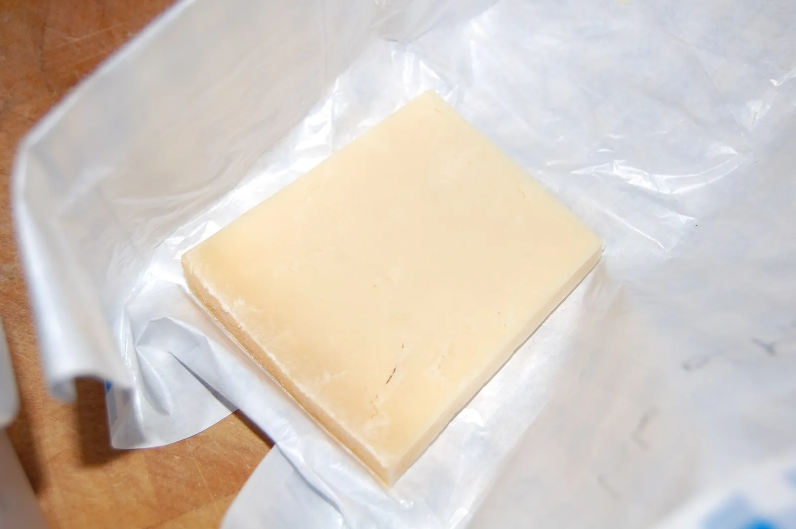 A pale yellow slab of cheddar cheese lying on crumpled clear plastic wrap on a wooden surface