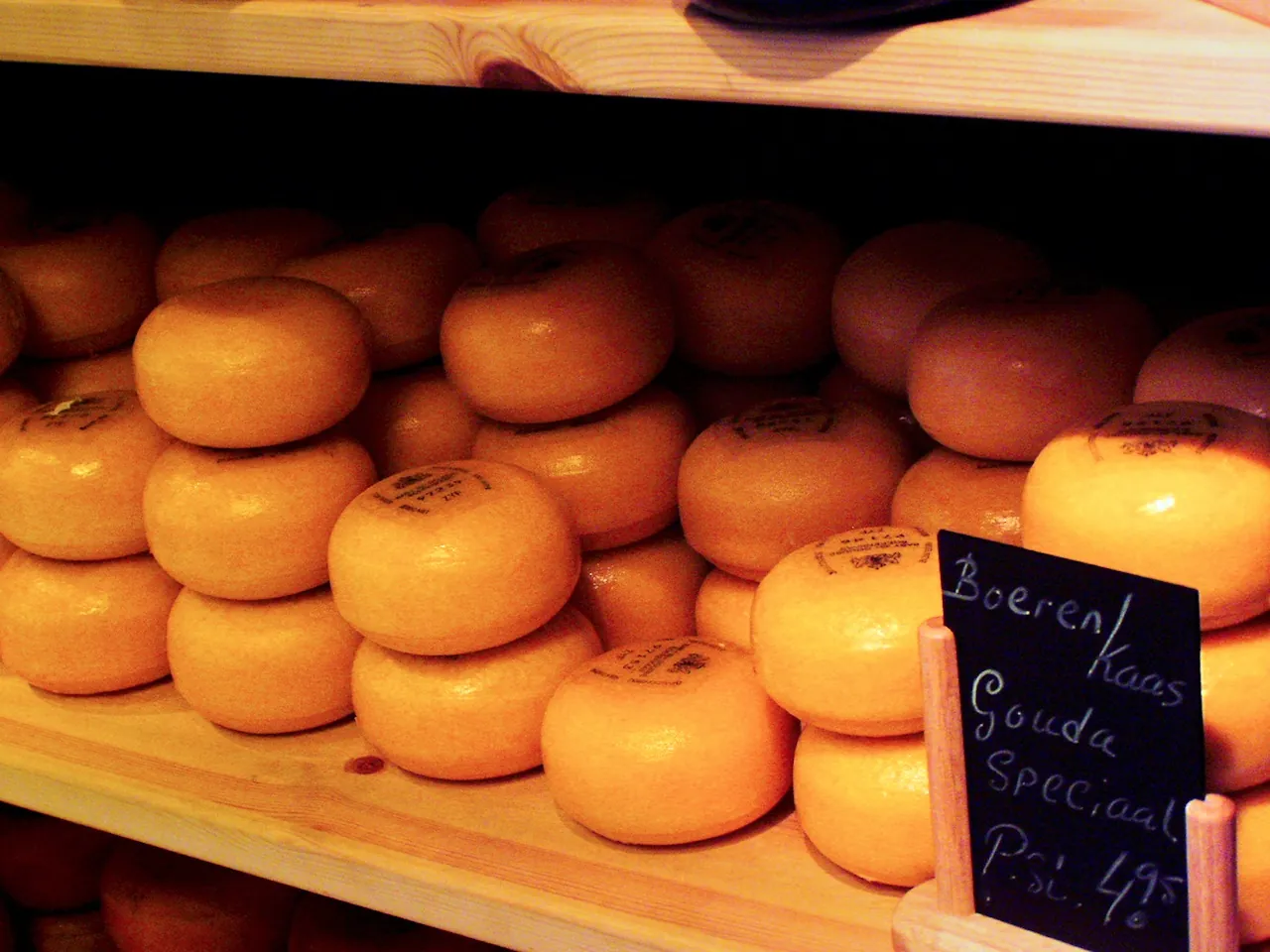 Rows of orange Gouda wheels on wooden shelves in an Amsterdam cheese shop, with a chalkboard sign