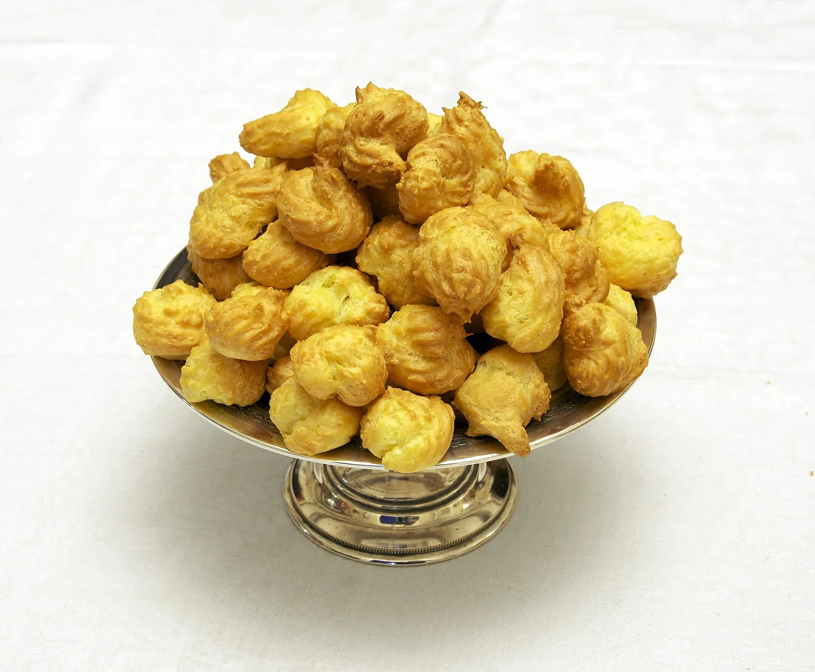 A silver pedestal dish heaped with small golden gougères, bite-sized cheese choux puffs, against a white background