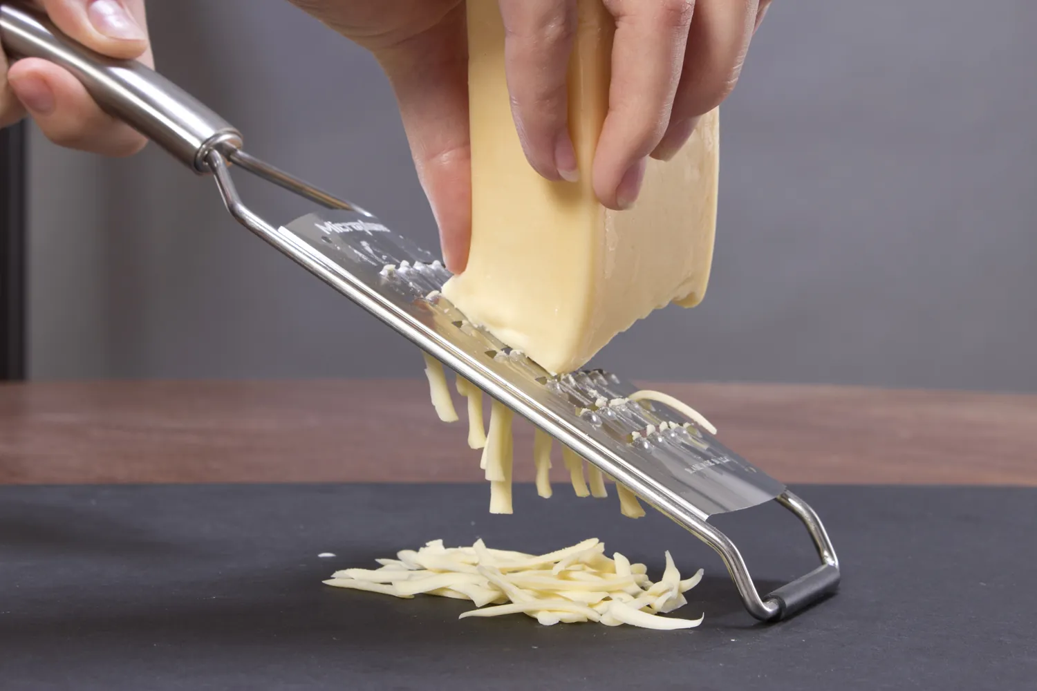 Hands grating a block of cheese on a handheld metal grater, with long shreds falling onto a dark counter below.