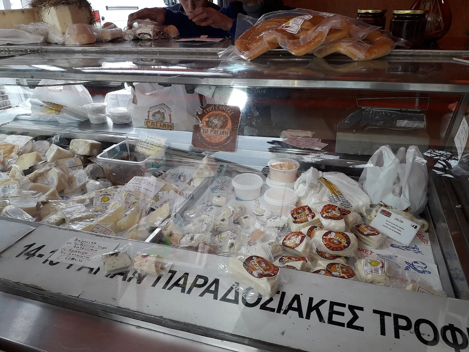 A glass deli counter in Athens holding white Greek cheeses, packed wedges and small round cheeses under a sign for traditional foods.