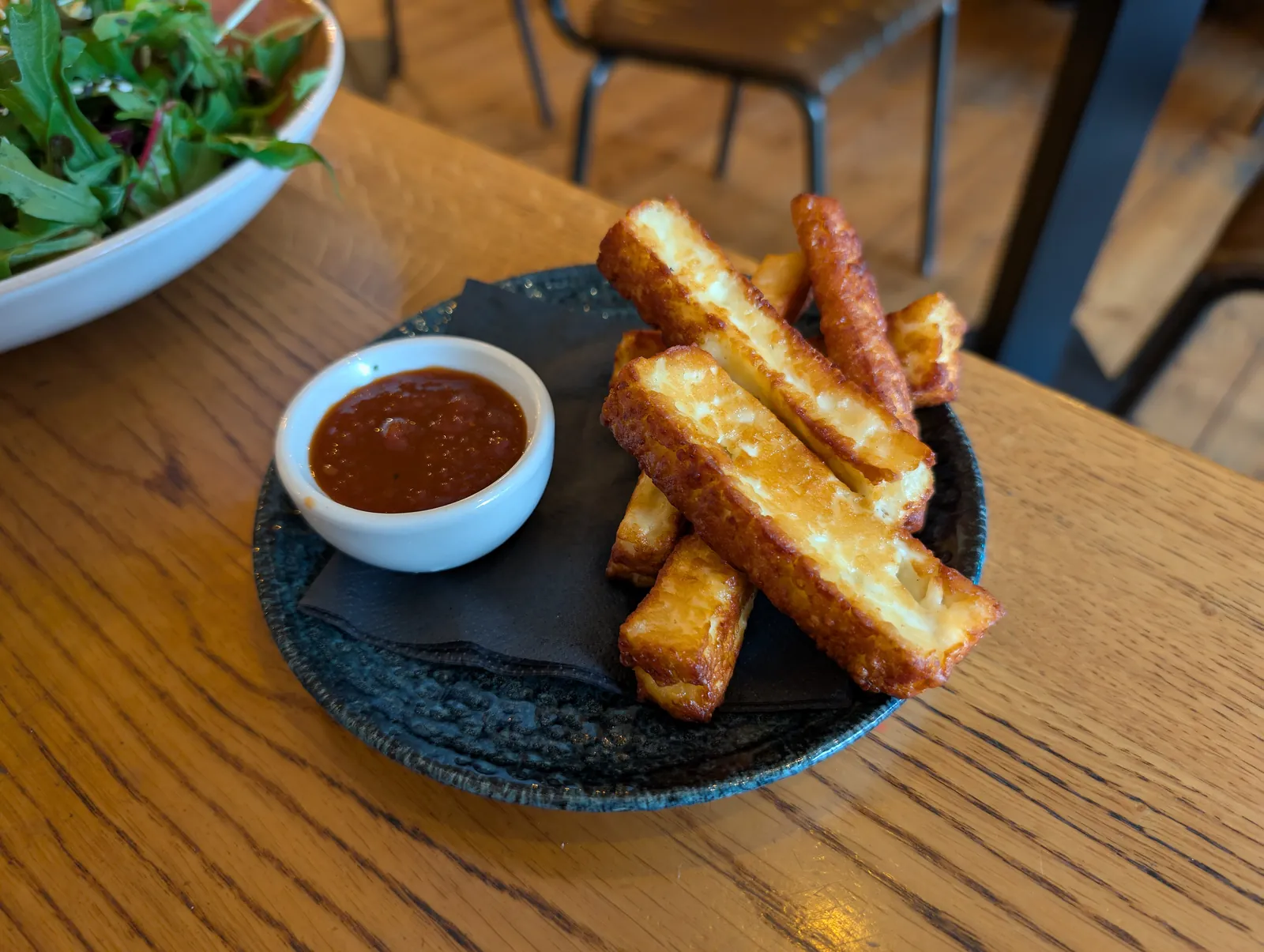 Golden fried halloumi sticks stacked on a dark plate beside a small bowl of red dipping sauce, with a green salad behind