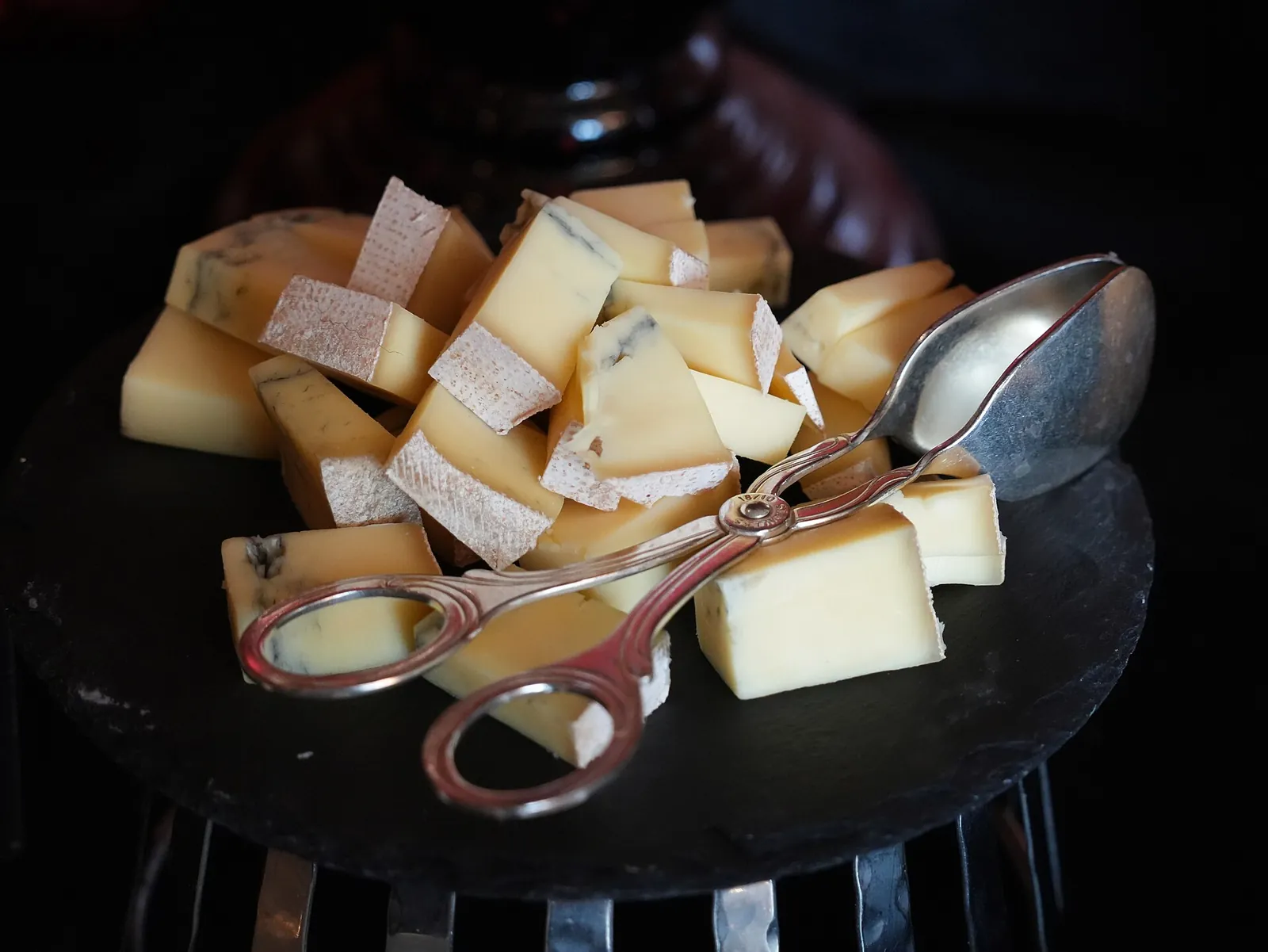 Cubes of aged cheese and a wedge of blue cheese piled on a black serving plate with a pair of metal tongs.