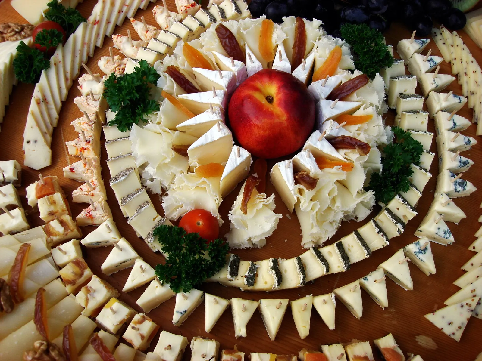 A large party platter of sliced cheeses arranged in rings, with frilly cheese rosettes and dried apricots around a nectarine