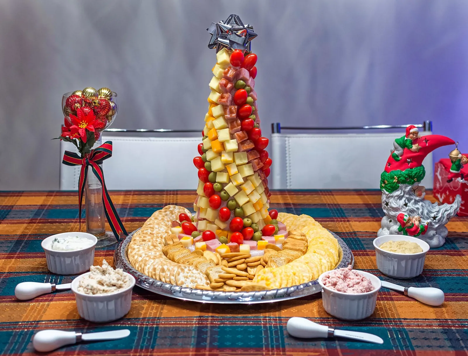 A Christmas tree made of cheese cubes, ham and tomatoes on a cracker platter, with dips, holiday figurines and a poinsettia.