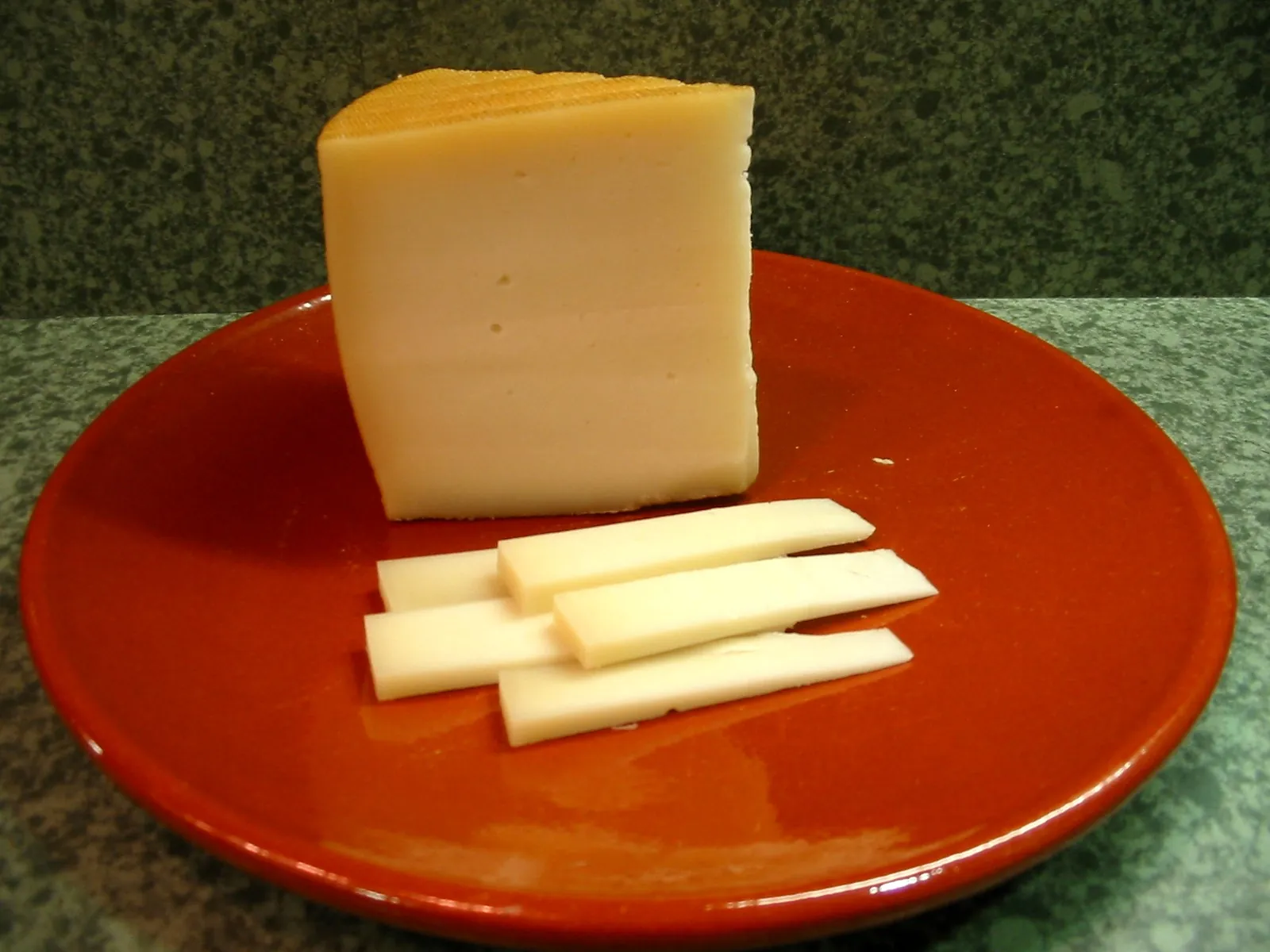 A wedge of Idiazabal sheep milk cheese on a red clay plate with thin slices beside it