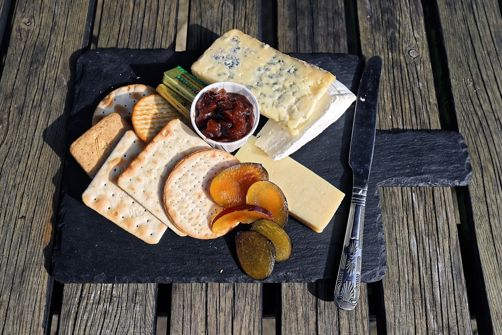 A slate board with blue cheese, crackers, sliced sweet potato chips and a small dish of cranberry sauce on a weathered wooden table.