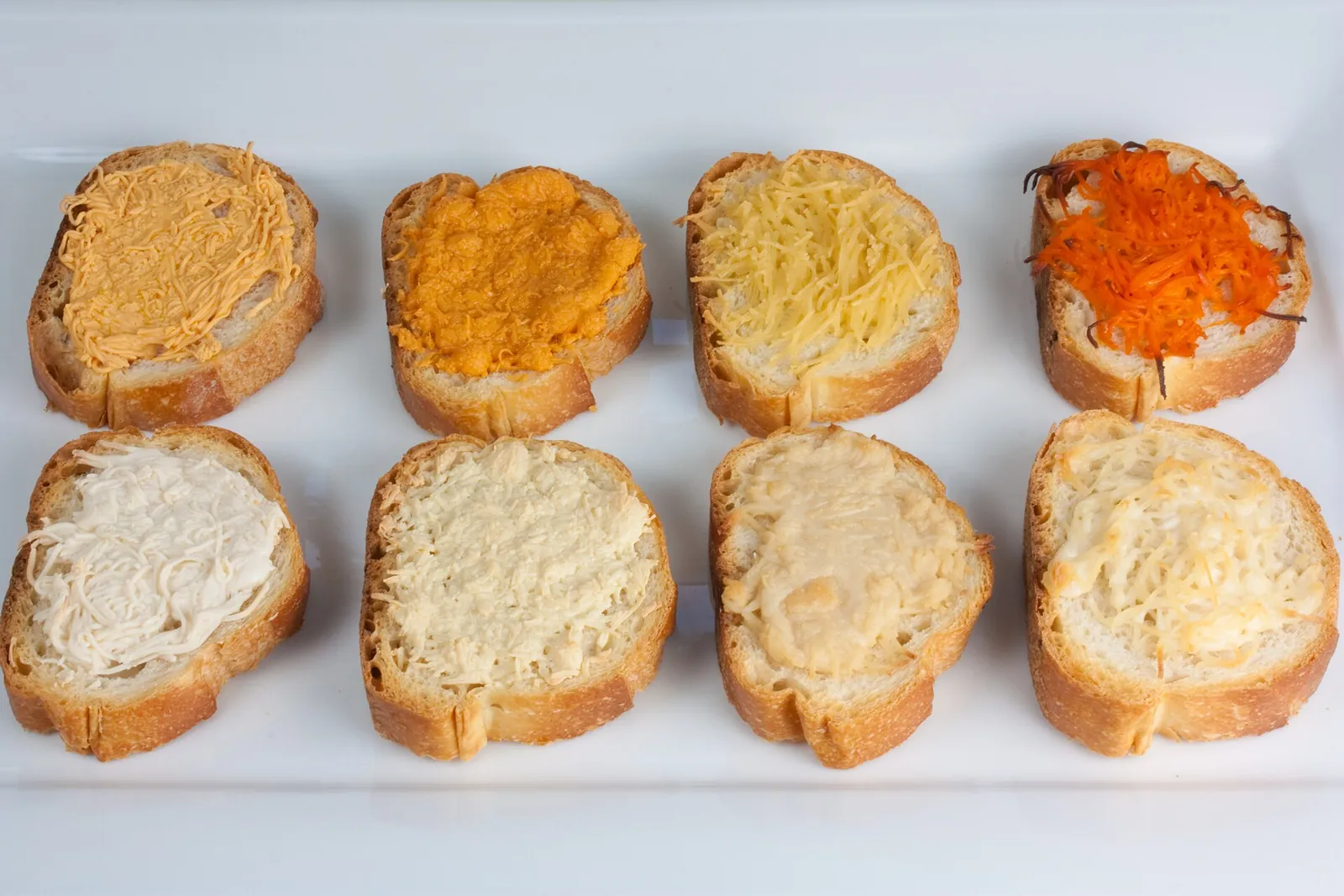 Eight slices of baguette topped with different plant based cheese spreads in white, cream, orange and red on a white tray.