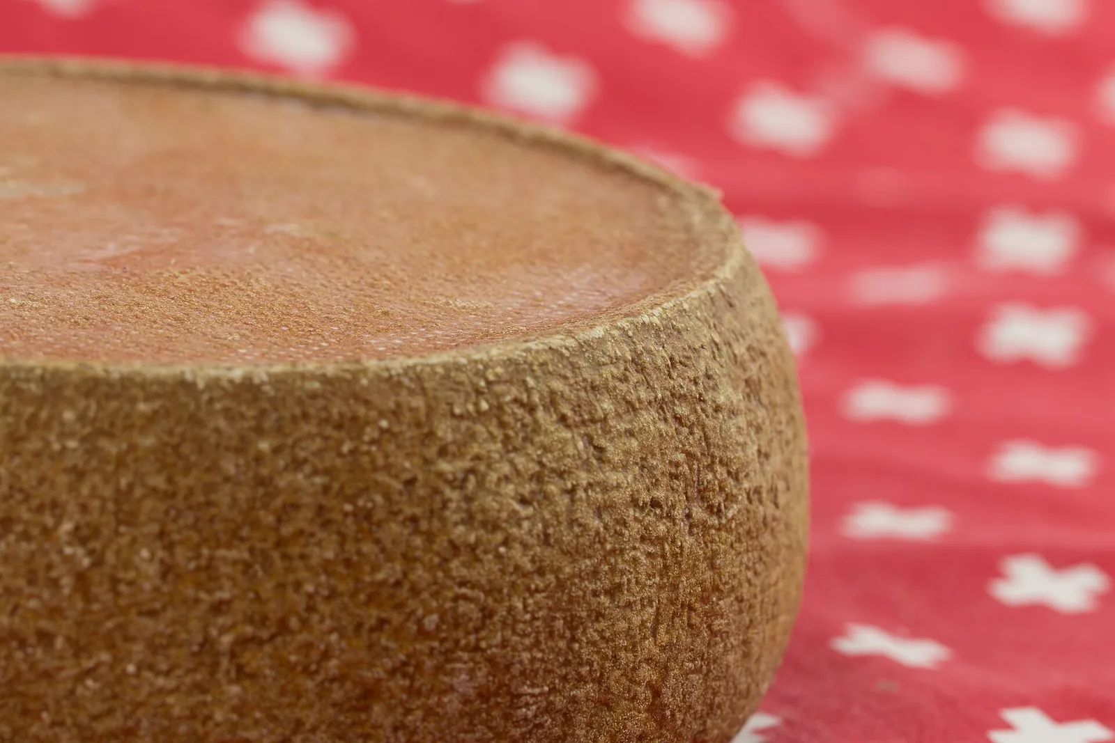 Close view of a round cheese showing its brown, finely textured natural rind and pale cut edge, on a red and white cloth