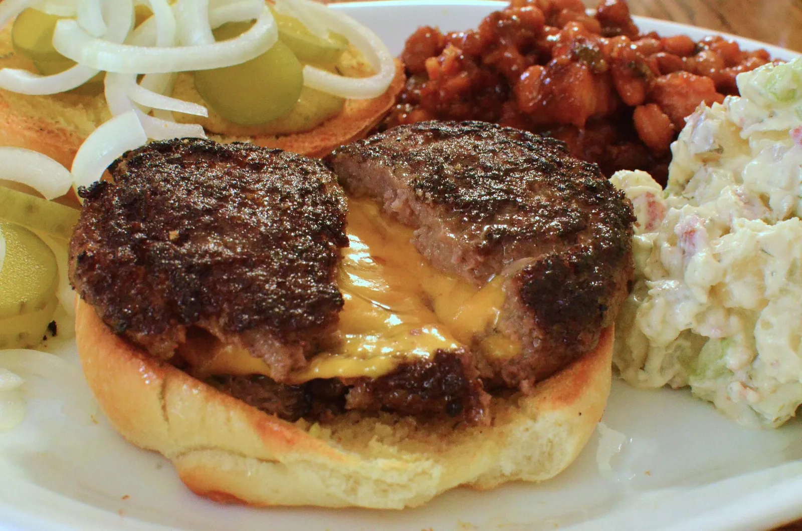 A thick griddled burger cut open so melted orange cheese runs out from the center, served with coleslaw and baked beans
