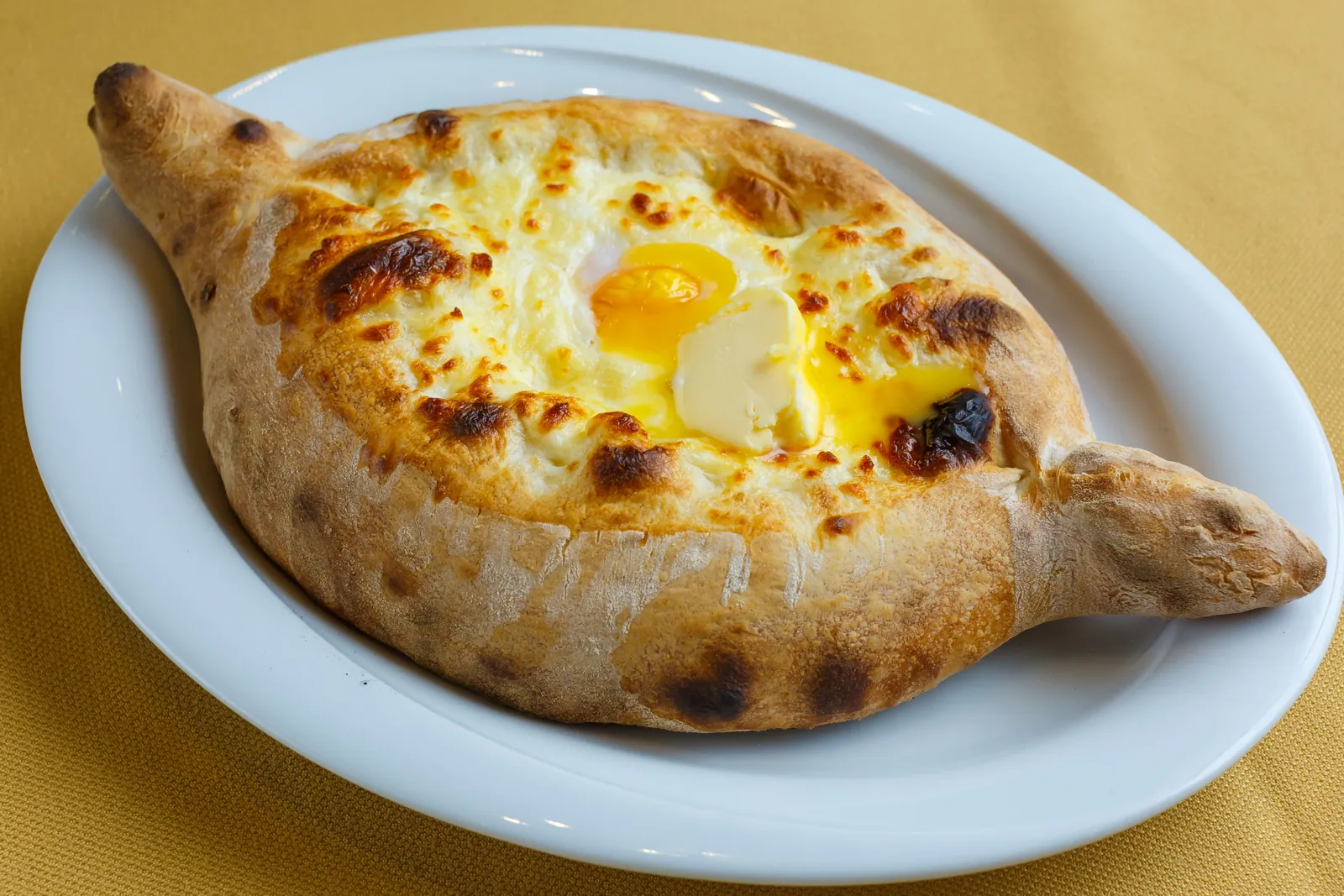 An Adjarian khachapuri, a boat-shaped bread baked golden and filled with melted cheese, a runny egg yolk and butter, on a white plate