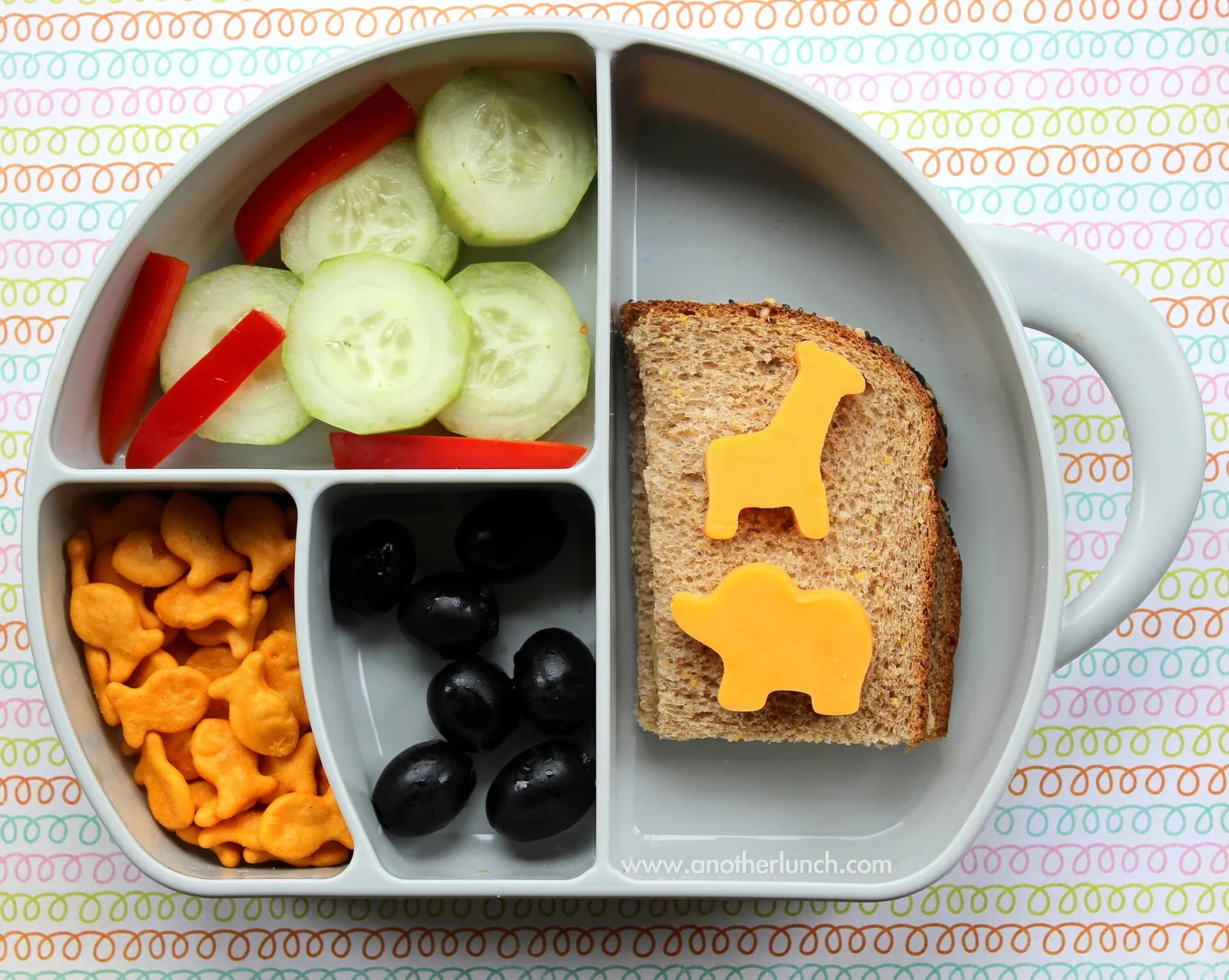 A preschool snack box with a sandwich topped with cheddar cut into animal shapes, cheese crackers, cucumber, red pepper and black olives