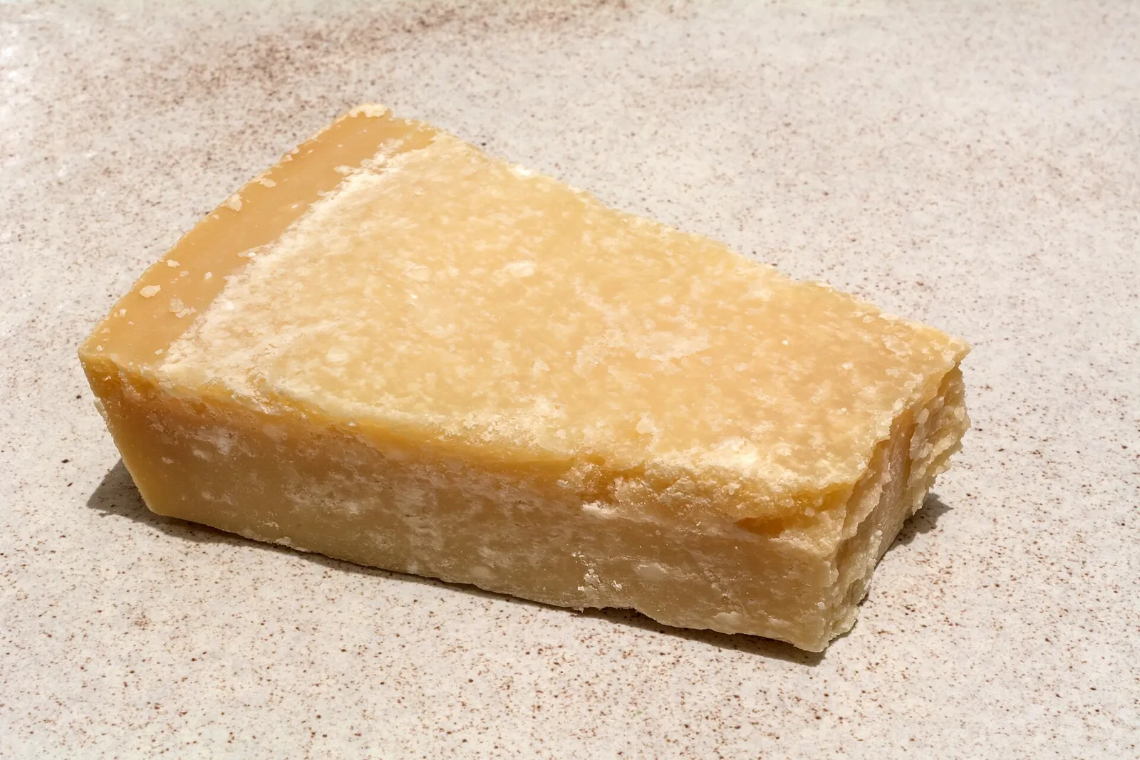 A rough chunk of aged Parmigiano Reggiano with a crumbly golden paste and speckled crystals, on a pale stone surface.