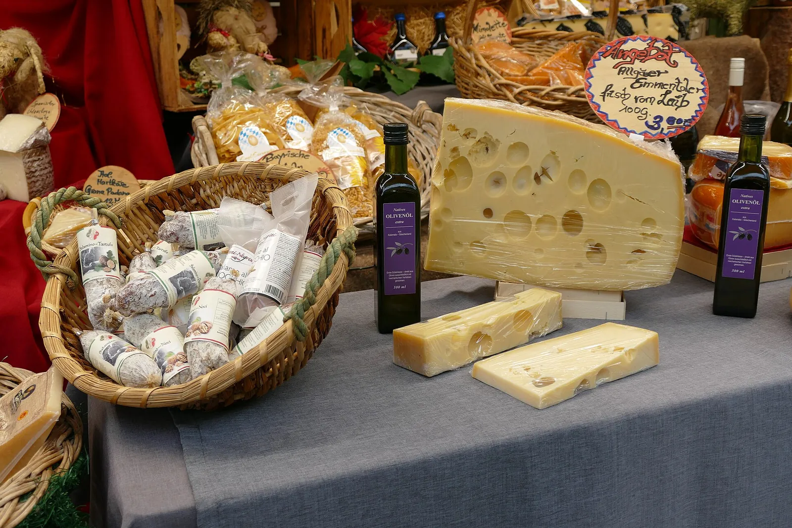A large wedge of Emmental with big round holes on a market stall table, beside smaller cut pieces, bottles and a basket of wrapped cheeses.