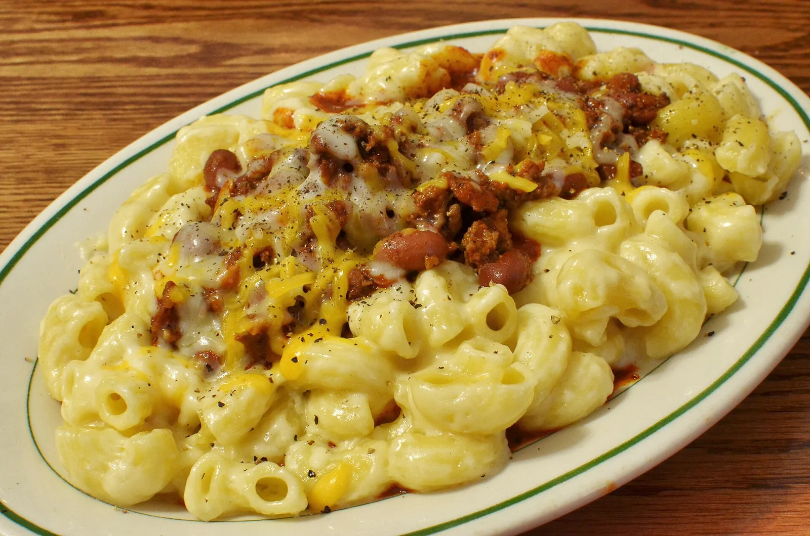 An oval plate of macaroni and cheese topped with chili, beans and shredded cheddar, the pasta pale and creamy underneath.