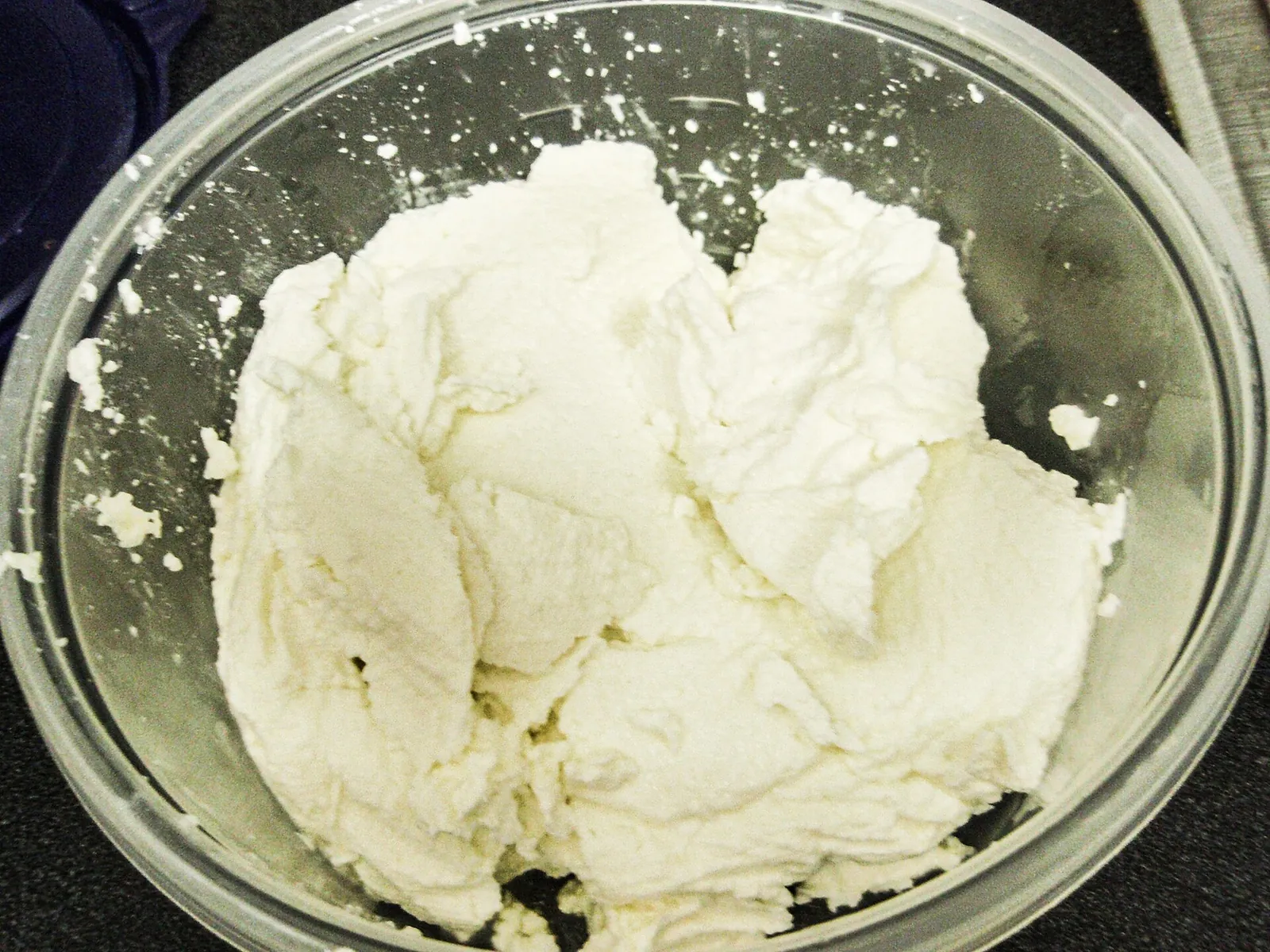 Soft, fluffy white fresh cheese heaped in a large clear mixing bowl, freshly drained and ready to be stirred smooth.