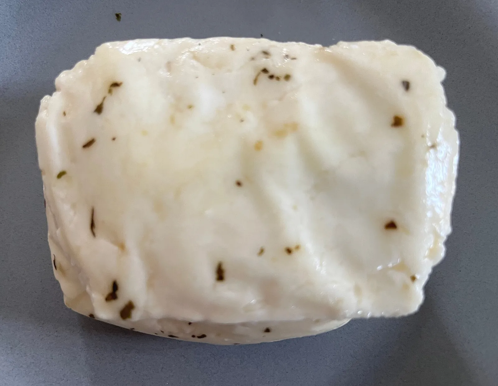 A whole block of fresh white halloumi flecked with dried mint, photographed from above on a gray surface.