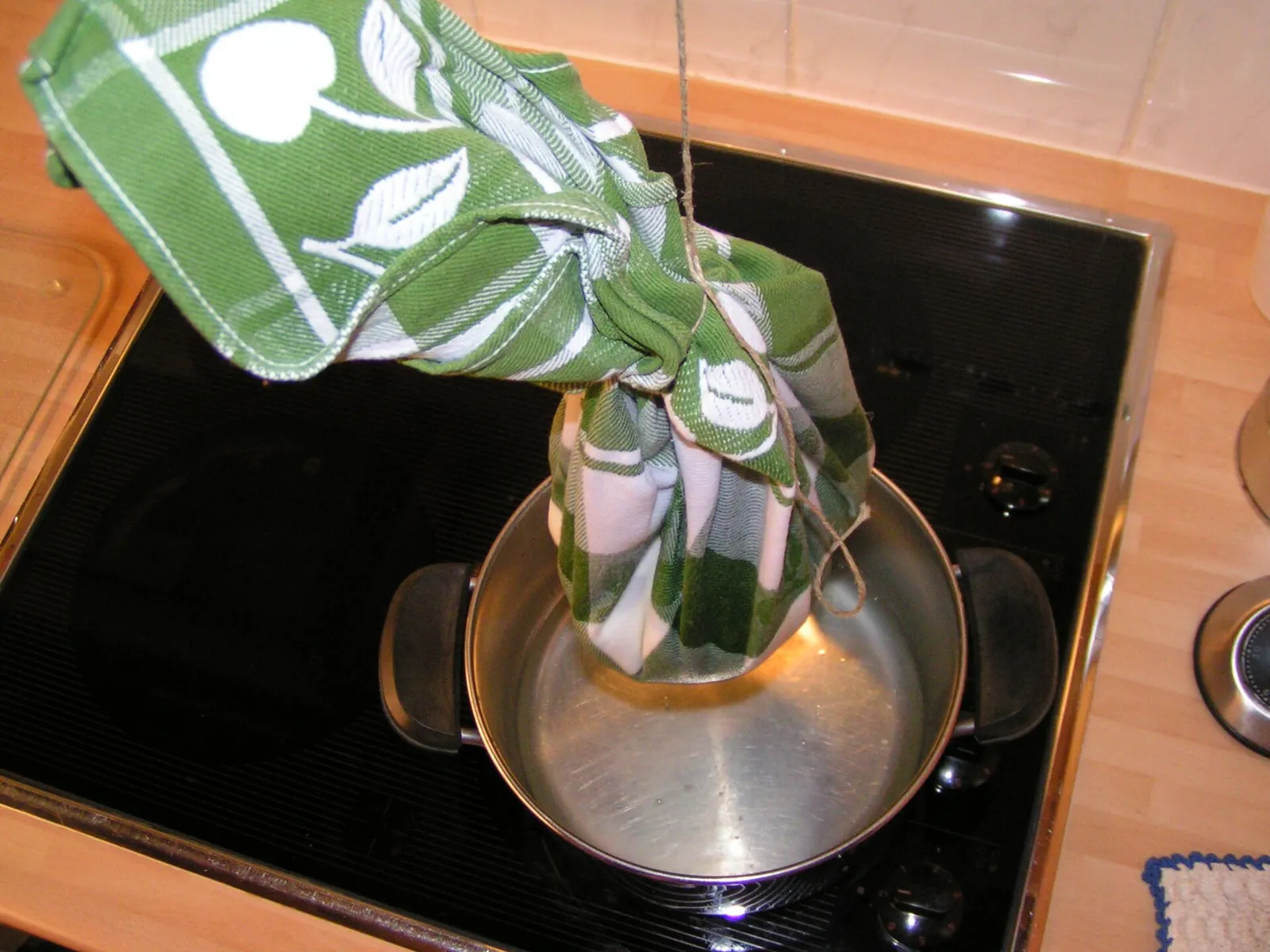 Strained yogurt tied up in a green kitchen towel, hanging over a steel pot on a stovetop to drain off the whey.