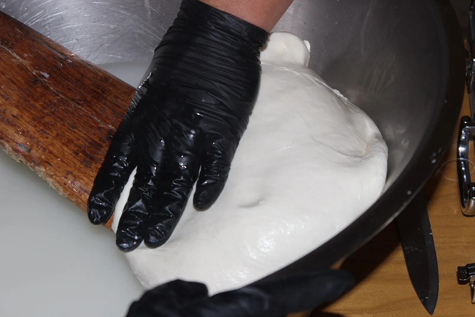 A gloved hand pressing and stretching a smooth mass of hot white curd in a large steel bowl.