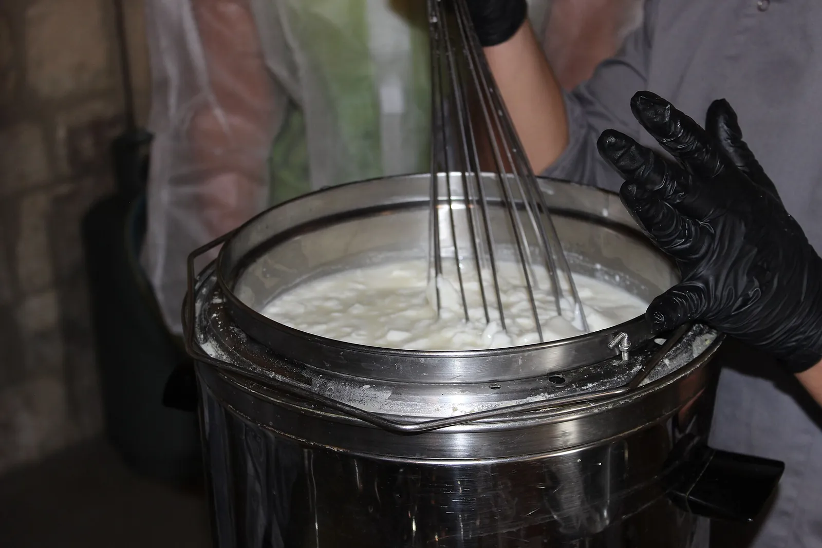 A cook in black gloves whisking a steel pot of hot milk where fine white ricotta curds are starting to form.