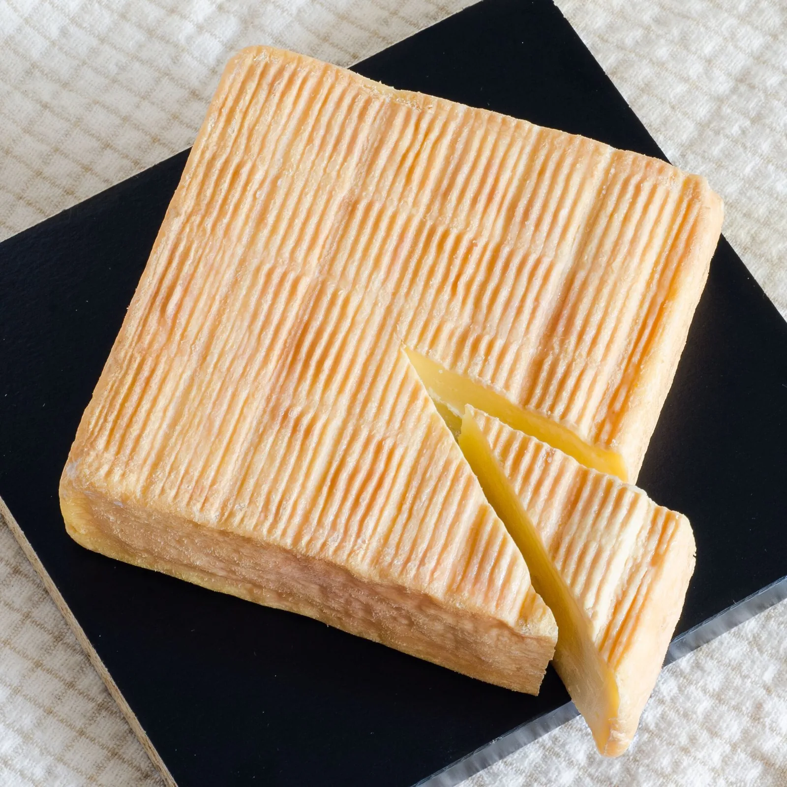 A square Maroilles with an orange washed rind and one wedge cut out, on a dark tray over a cloth