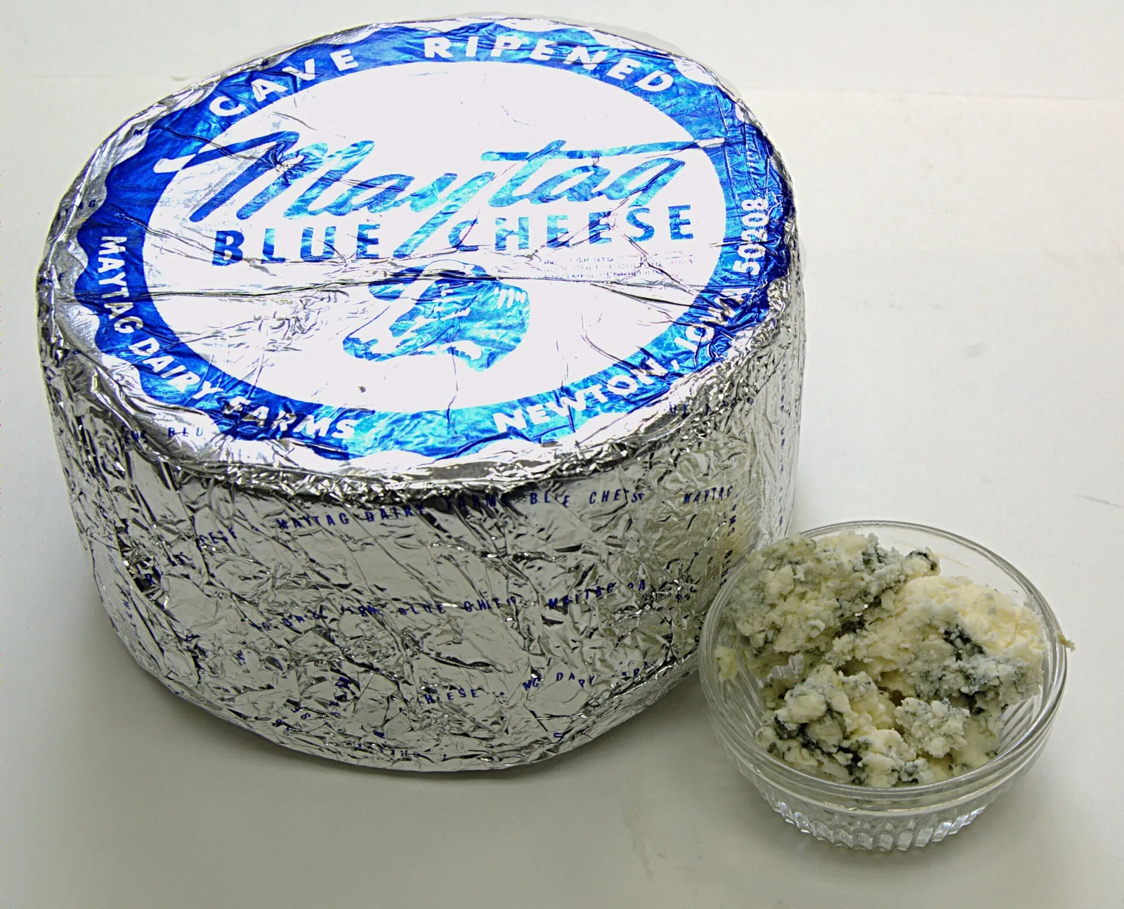 A foil-wrapped wheel of Maytag Blue Cheese beside a dish of blue cheese crumbles