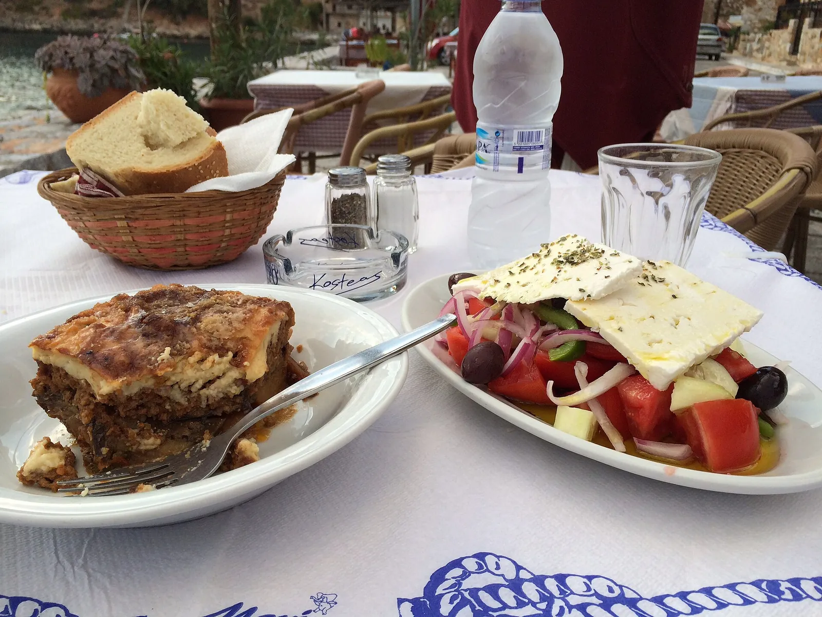 A taverna table set with baked moussaka and a Greek salad of tomato, olives and feta, beside a basket of bread.