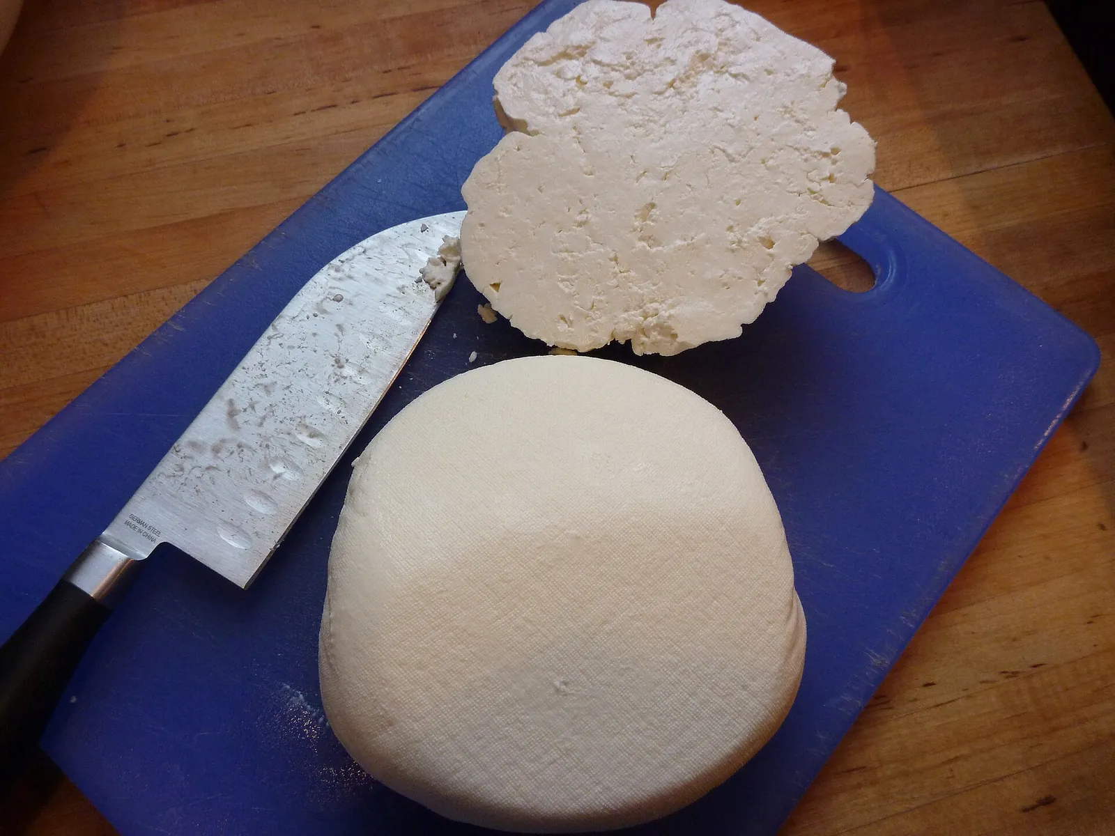 A fresh Mizithra wheel and a cut slice on a blue cutting board beside a chef's knife