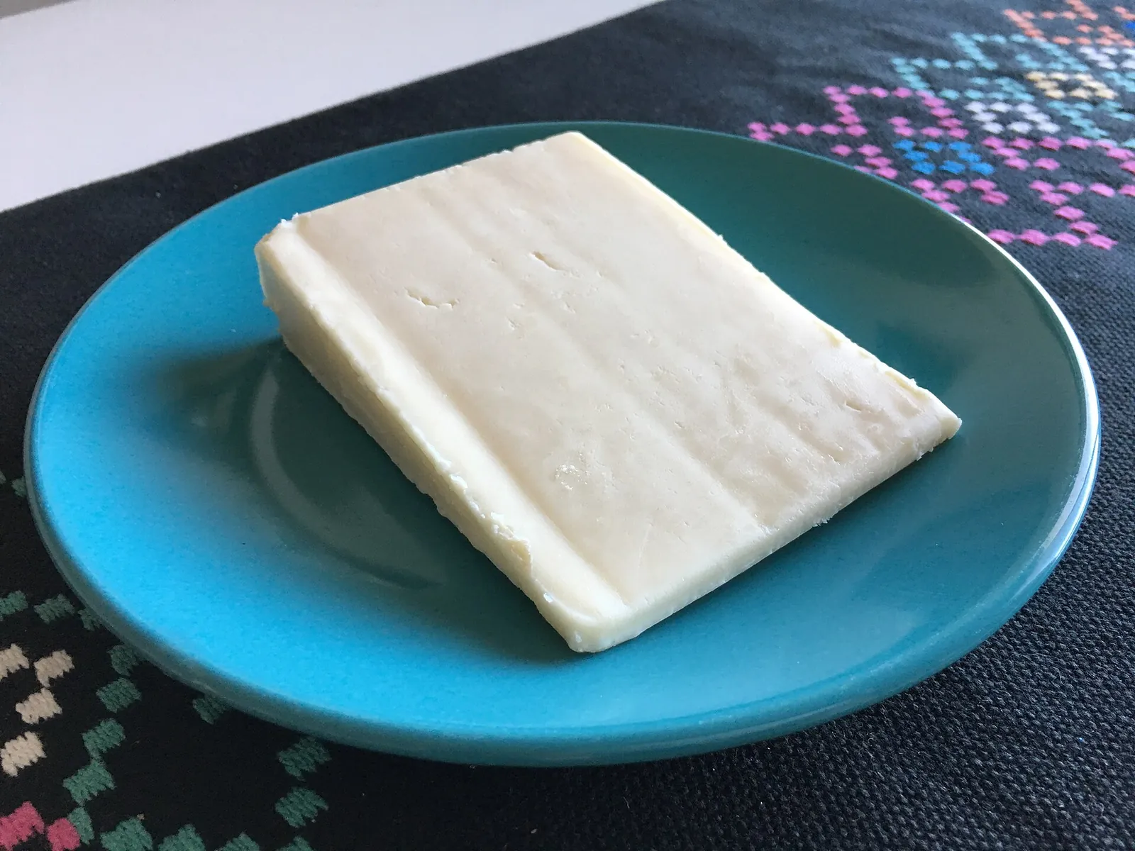 Pale rectangular wedge of semi soft Sonoma Jack cheese resting on a teal plate over a patterned black cloth.