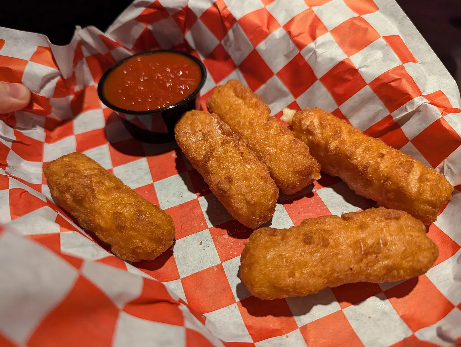 Golden fried mozzarella sticks in a basket lined with red and white checkered paper, with a small cup of marinara for dipping.