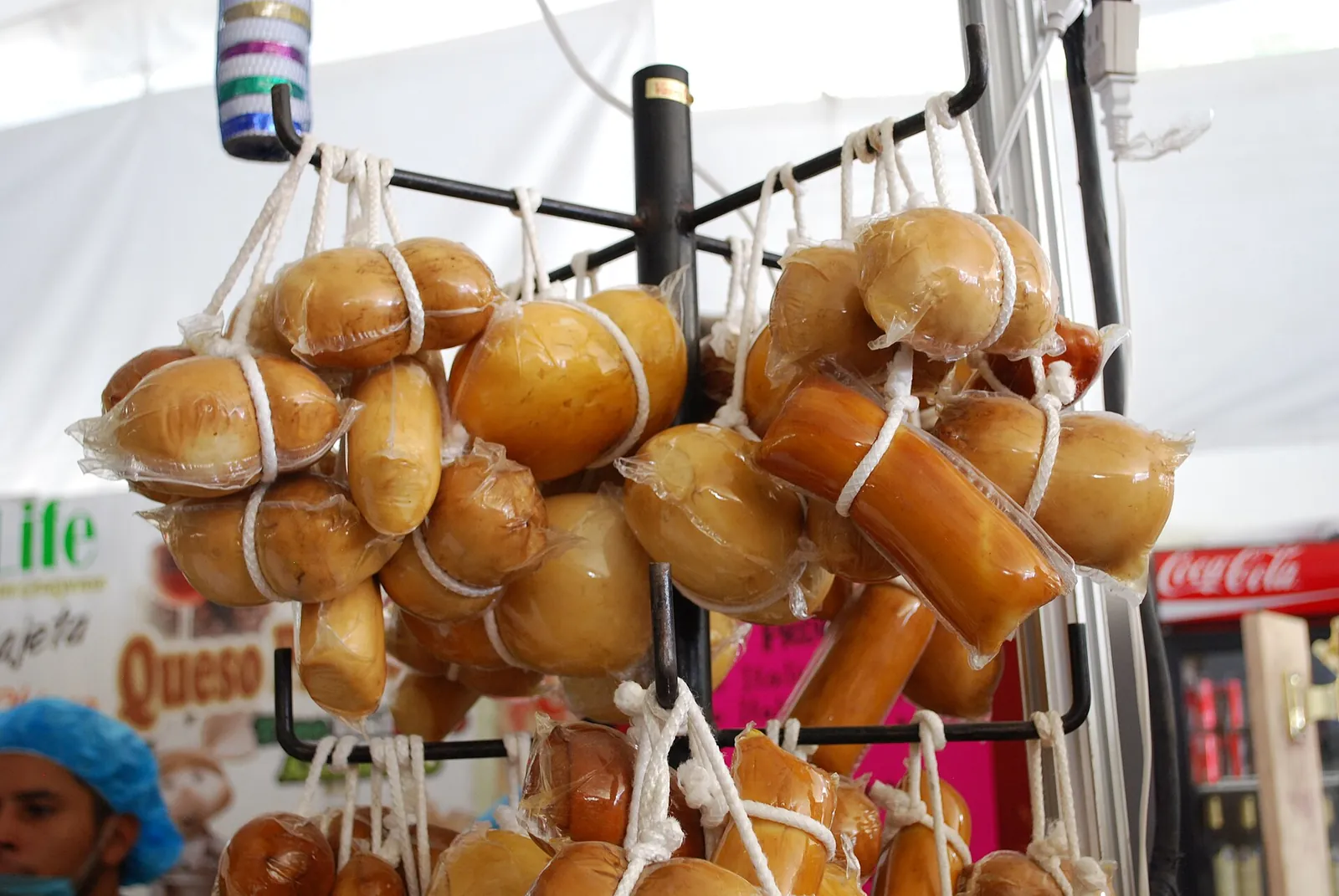 Dozens of smoked, string tied provolone style cheeses in plastic wrap hanging from a market stall rack.