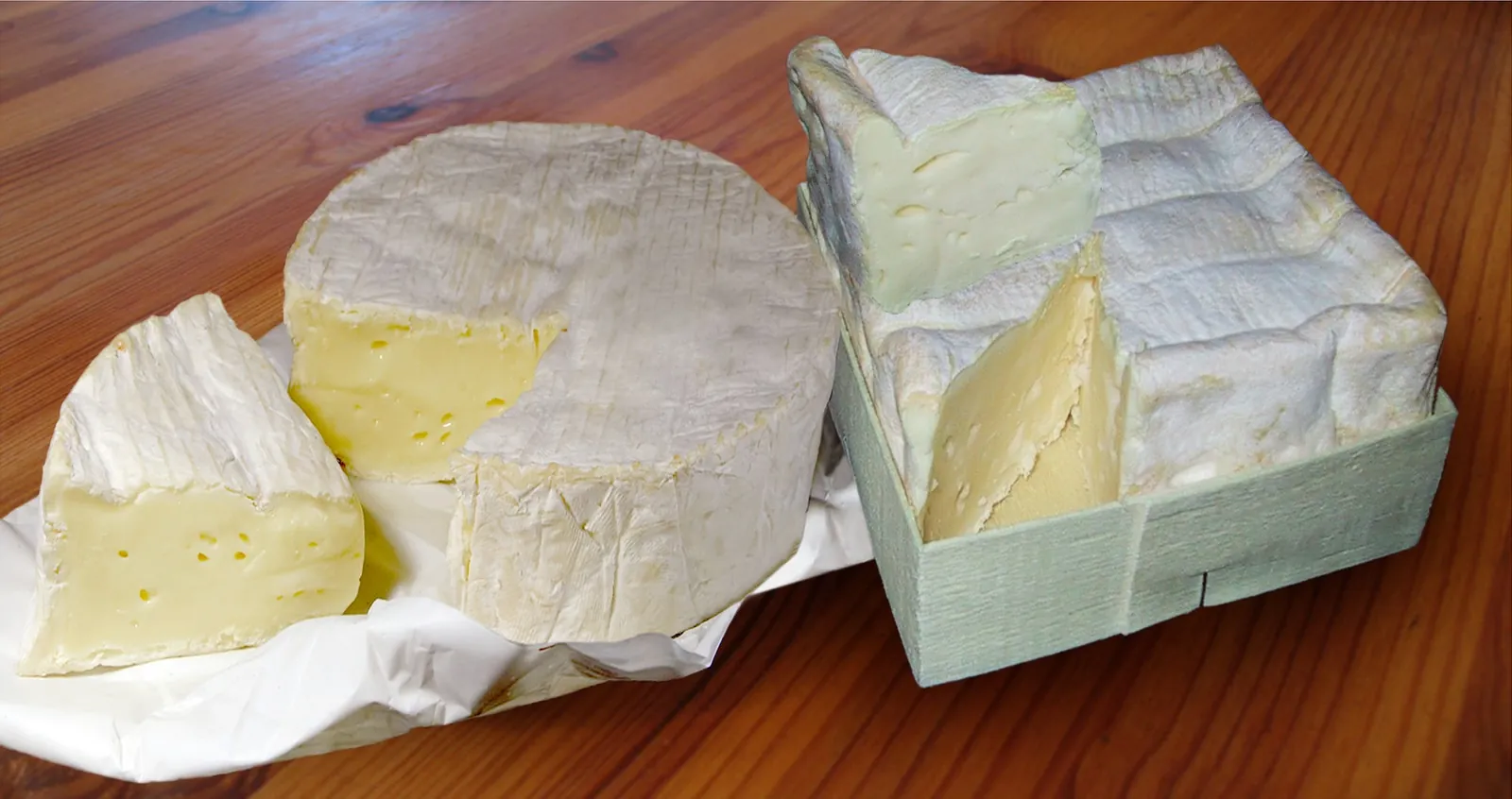 A whole Camembert with a wedge cut out beside a Pont-l'Évêque in its square wooden box, on white paper over a wooden table.