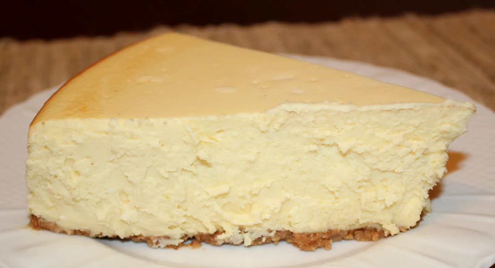 A tall wedge of New York style baked cheesecake on a white plate, showing the dense pale filling above a thin graham cracker crust
